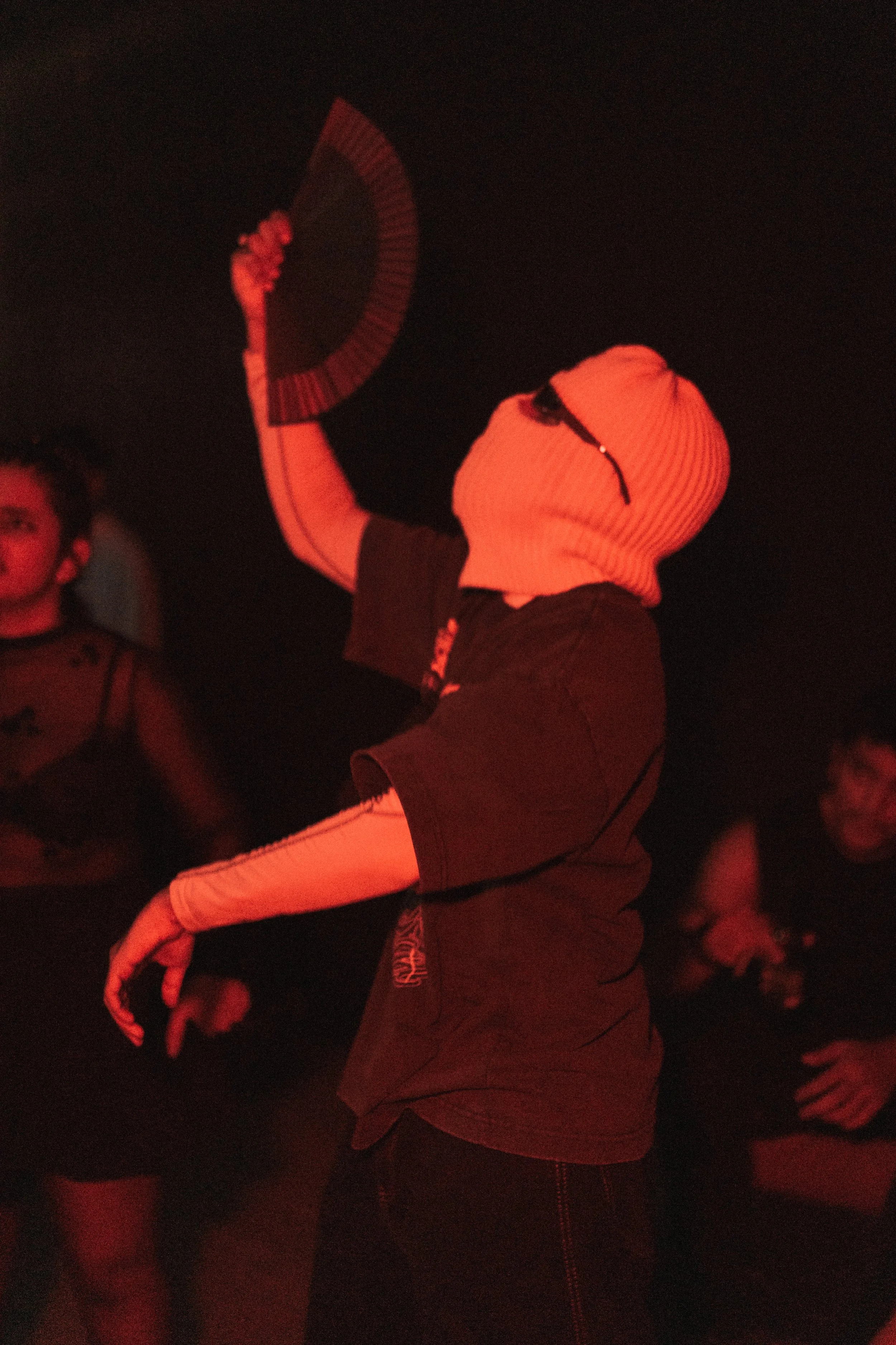 Person wearing a white ski mask, black t-shirt, and glasses, holding a fan, surrounded by other people in dark setting with red lighting.