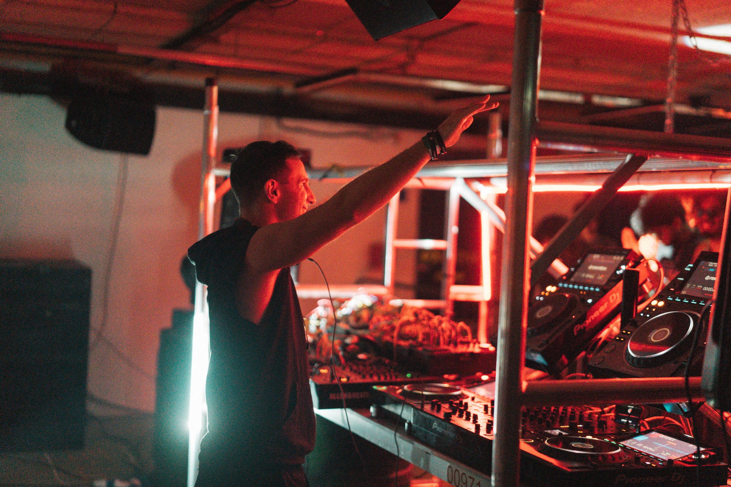 A DJ is performing at a music event, standing behind a DJ mixer with various equipment, under red lighting, with other people in the background.