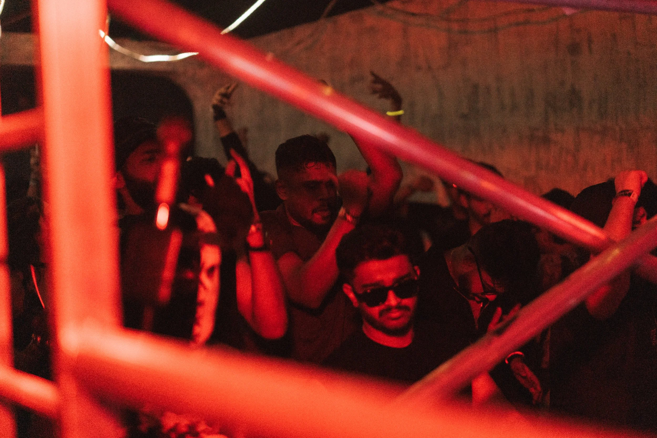 People attending a concert or event at night, with some wearing sunglasses, in a dimly lit environment with red lighting, seen through orange metal bars.
