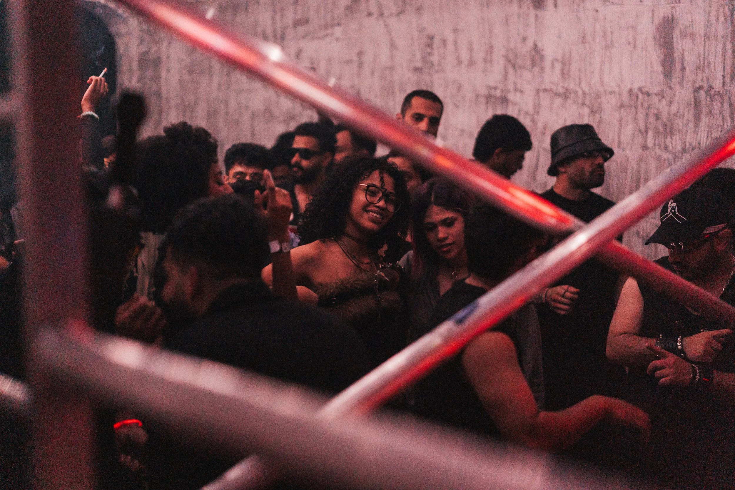 A group of people at a party or nightclub, with some dancing and socializing, with a pinkish lighting and a plain wall in the background.
