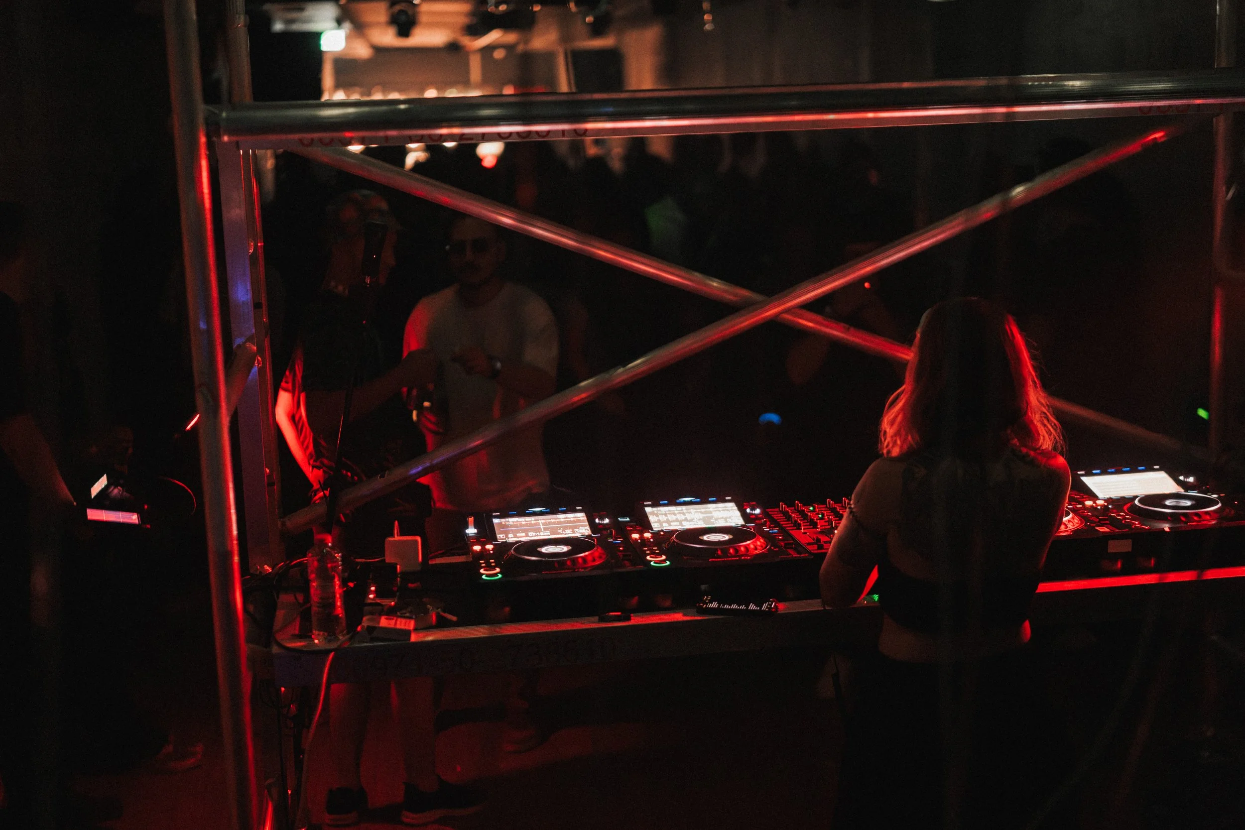 A DJ woman with blonde hair performing in a dimly lit nightclub with red lighting, as people dance in the background.