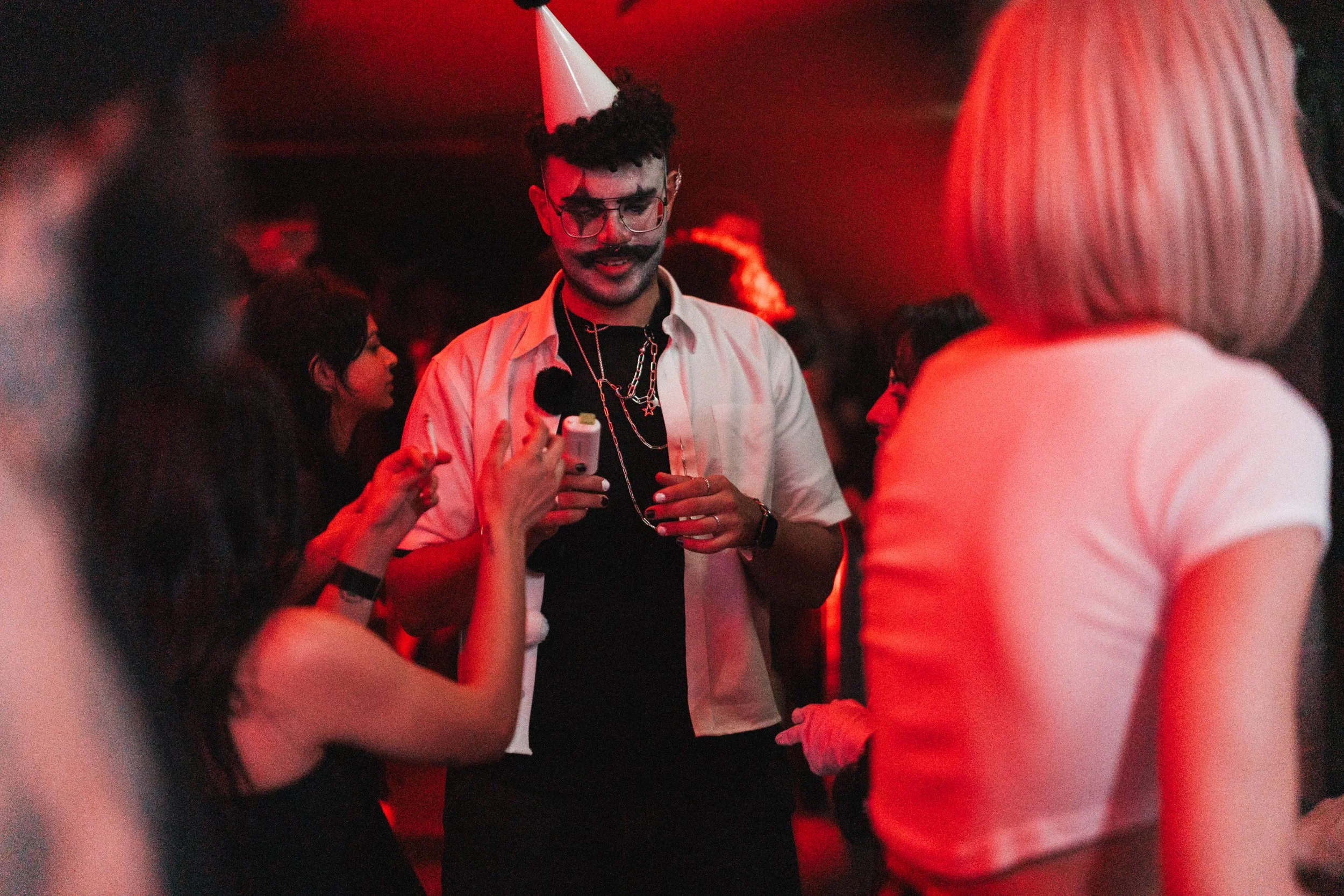 A group of young people celebrating at a party with red lighting. One person is wearing a party hat and sunglasses, holding a video camera. Others are engaged in conversation and dancing.