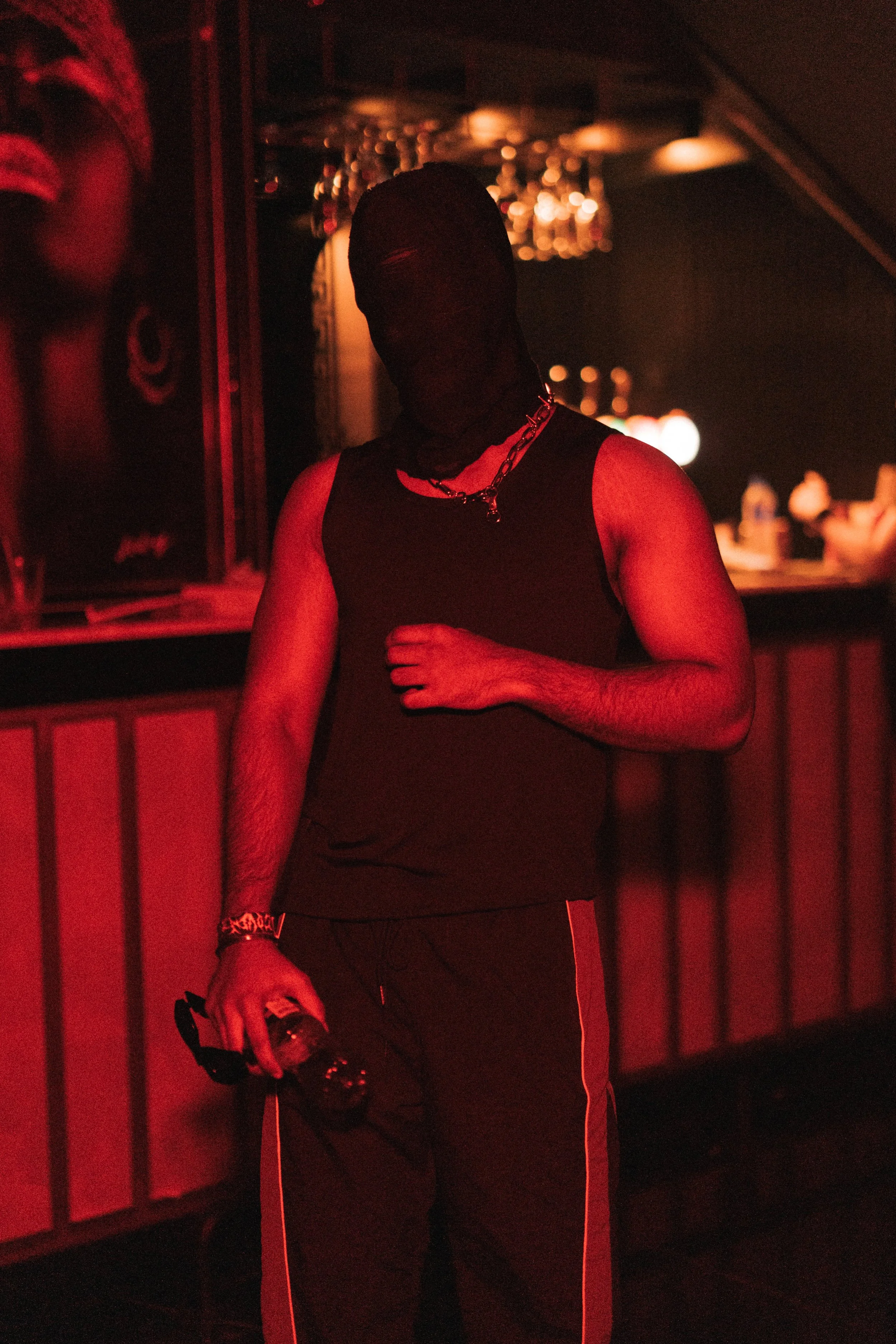 A man dressed in black clothing with a chain necklace is standing in a dimly lit bar or nightclub with red lighting. He is holding sunglasses and a drink, and a mask covers his face.