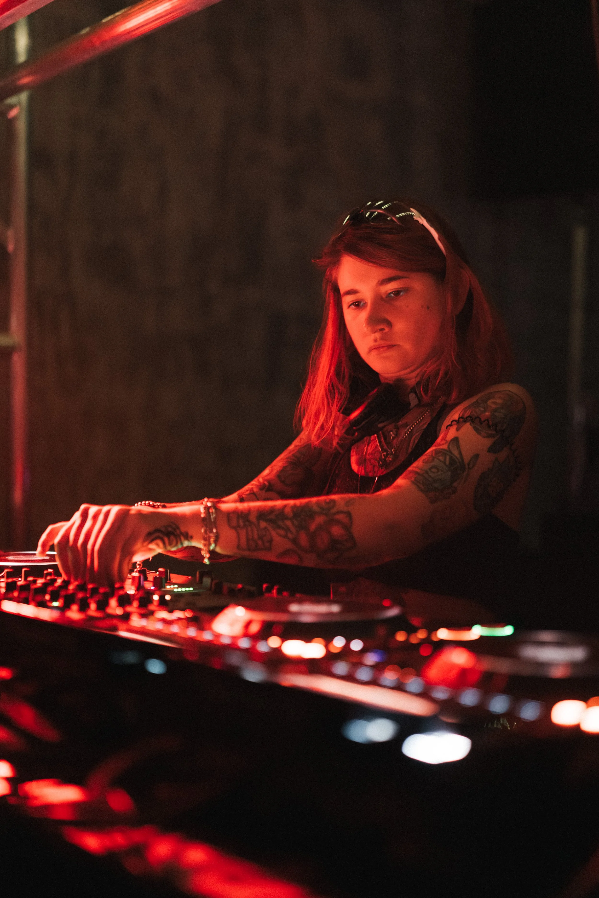 A female DJ with red hair and tattoos on her arms, wearing a headband and headphones around her neck, operating a DJ mixer in a dimly lit environment with red lighting.