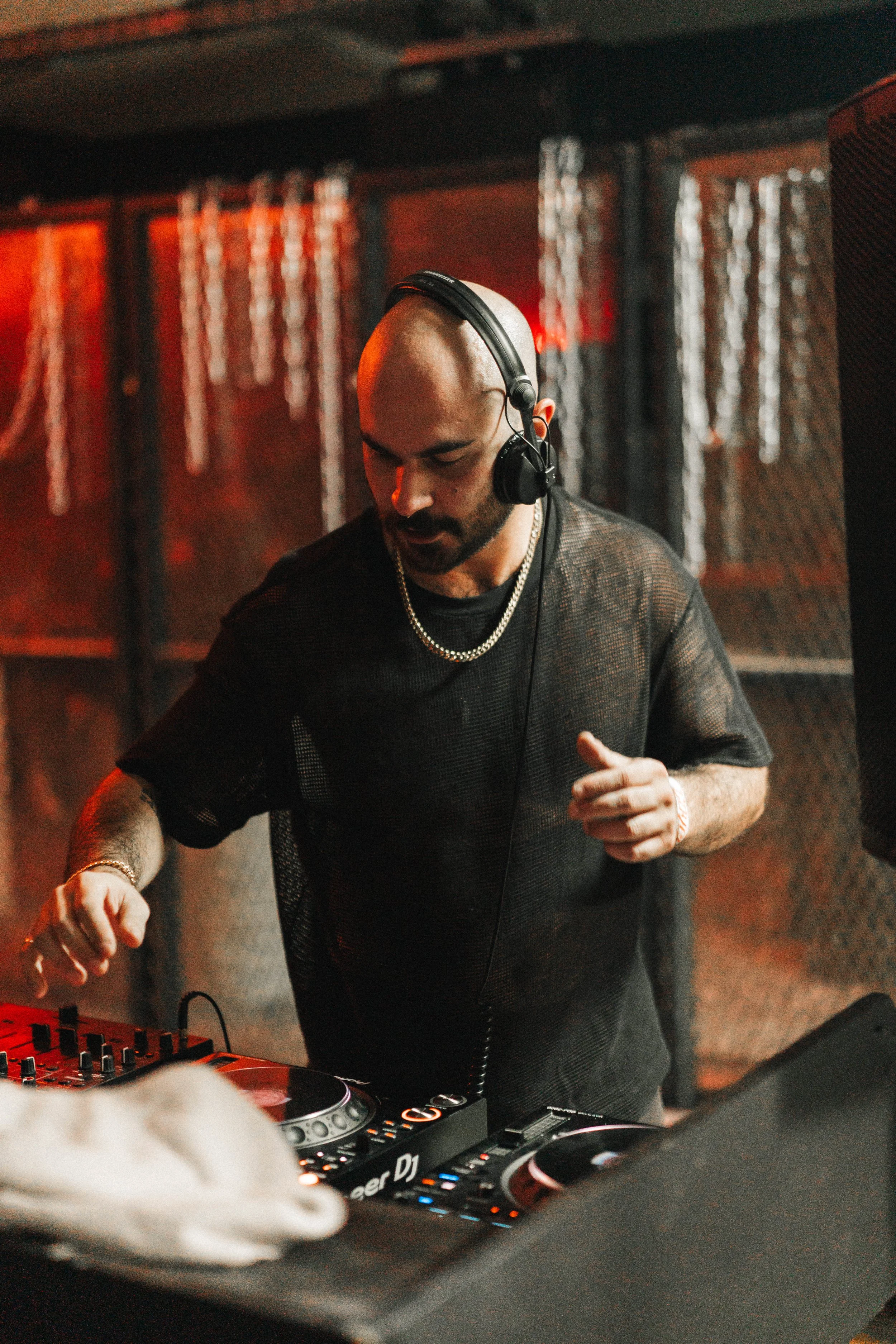 A DJ with a shaved head and beard wearing headphones, a black mesh shirt, and jewelry, operating DJ equipment in a club with red lighting.