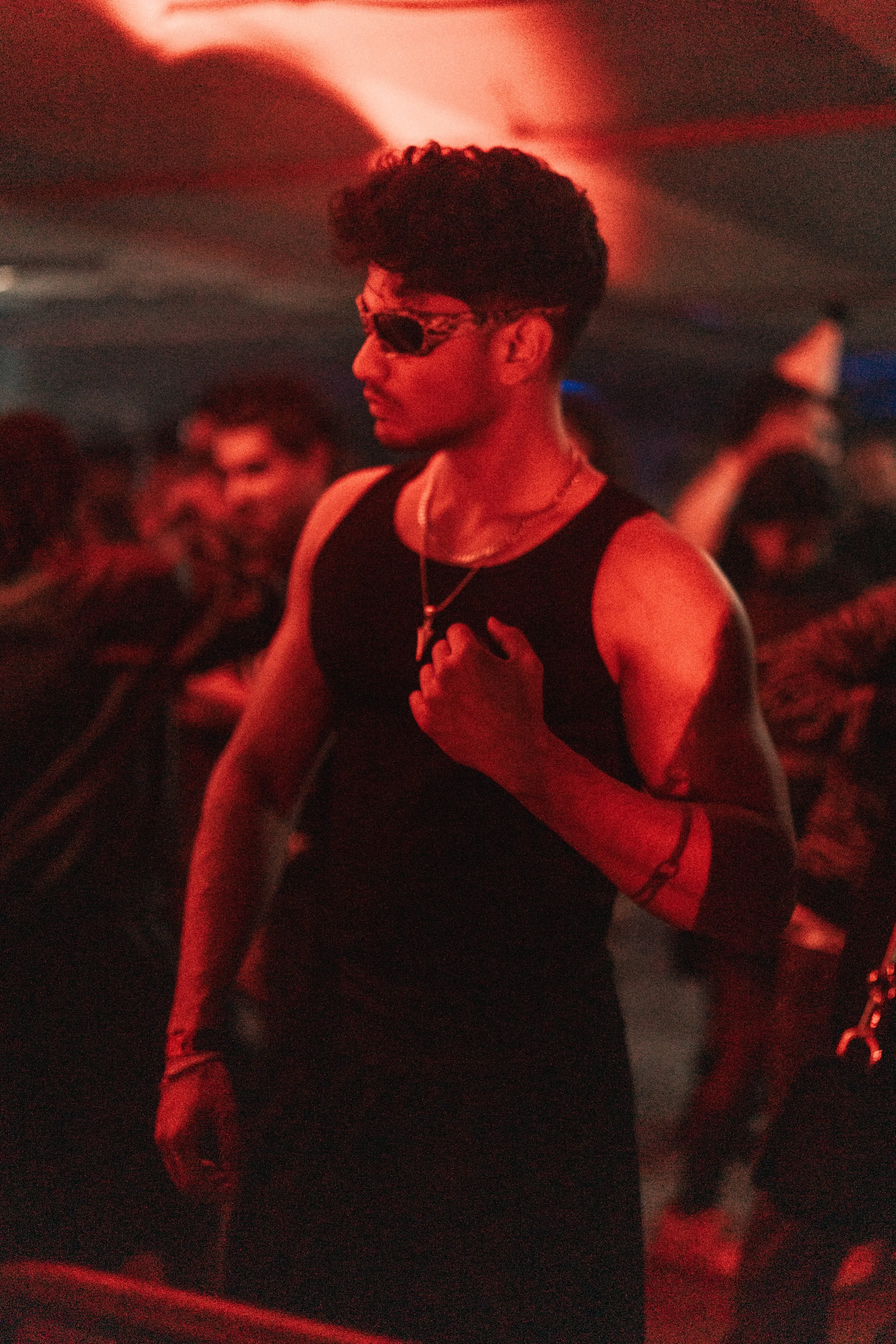 A young man wearing sunglasses and a black sleeveless shirt, with tattoos on his arms, standing in a dimly lit environment with red lighting, surrounded by other people.