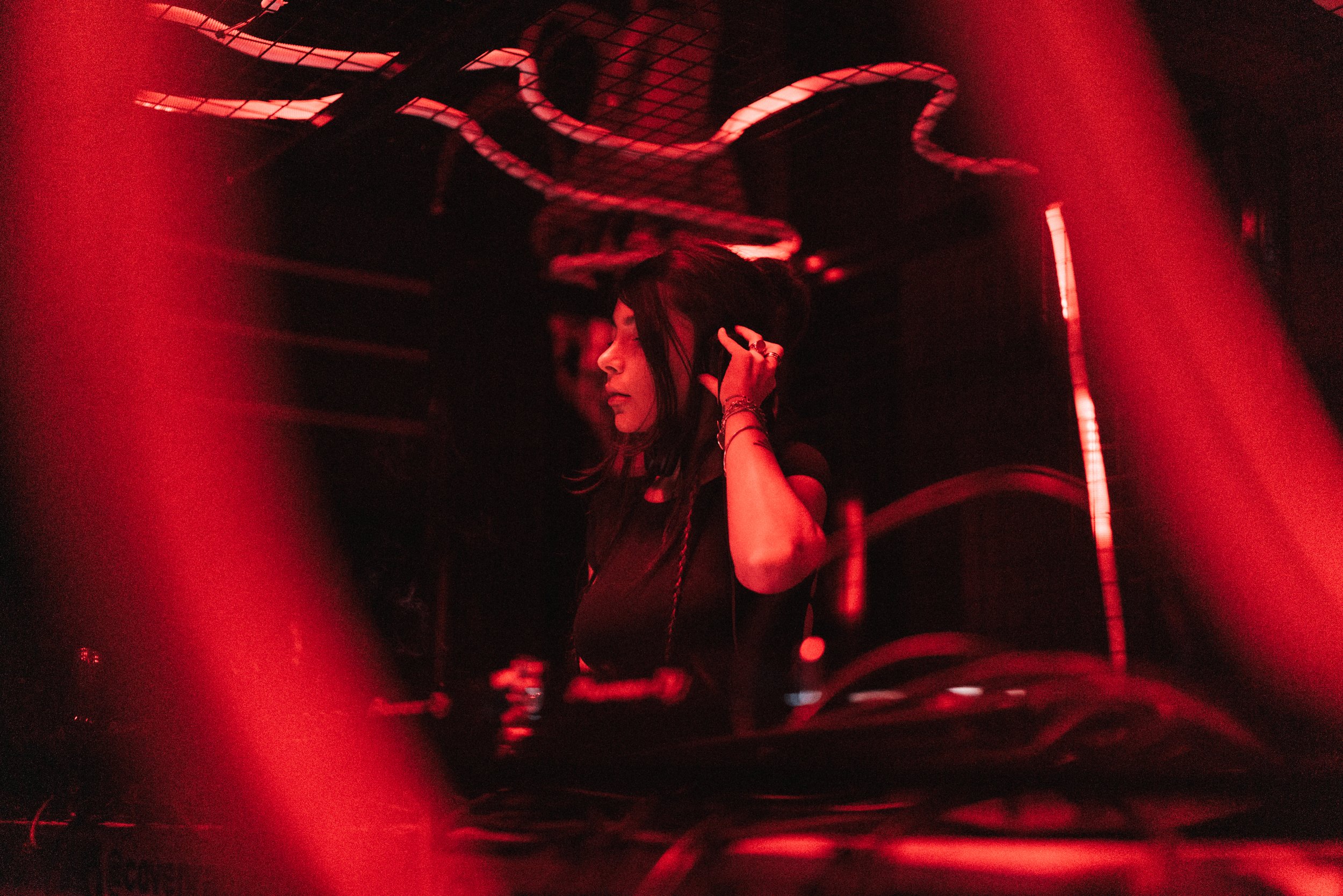 A woman DJing with red lighting in a club or concert setting, adjusting her headphones.
