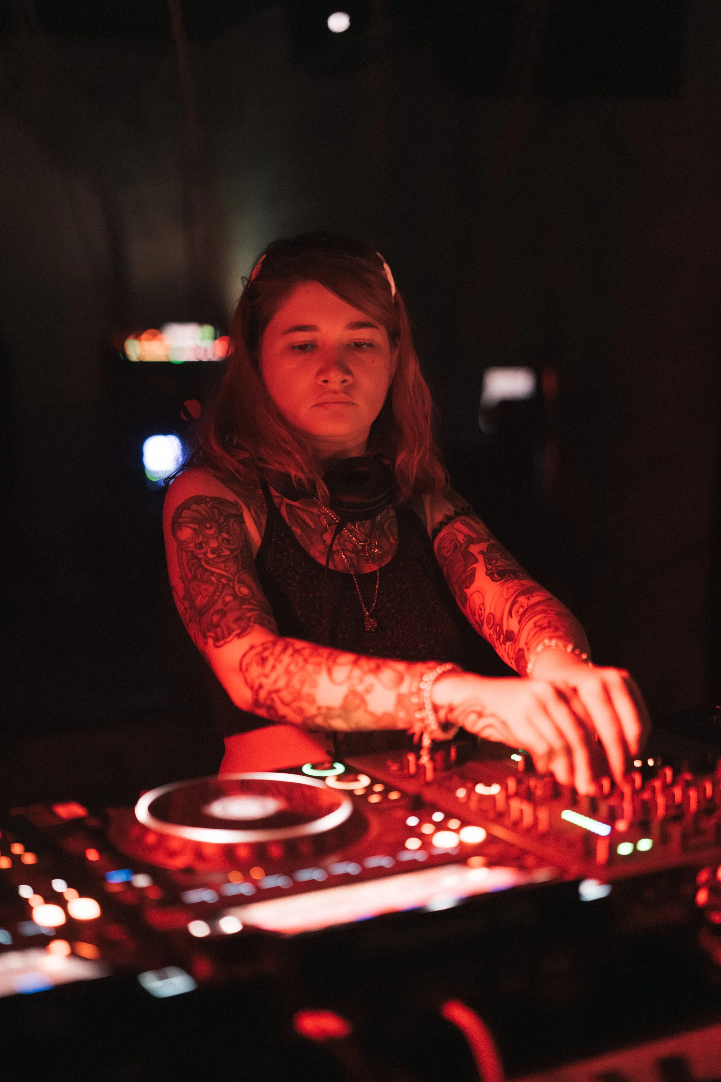 A female DJ with tattoos on her arms performing at a dimly lit event, illuminated by red lighting, with her hands on DJ equipment.