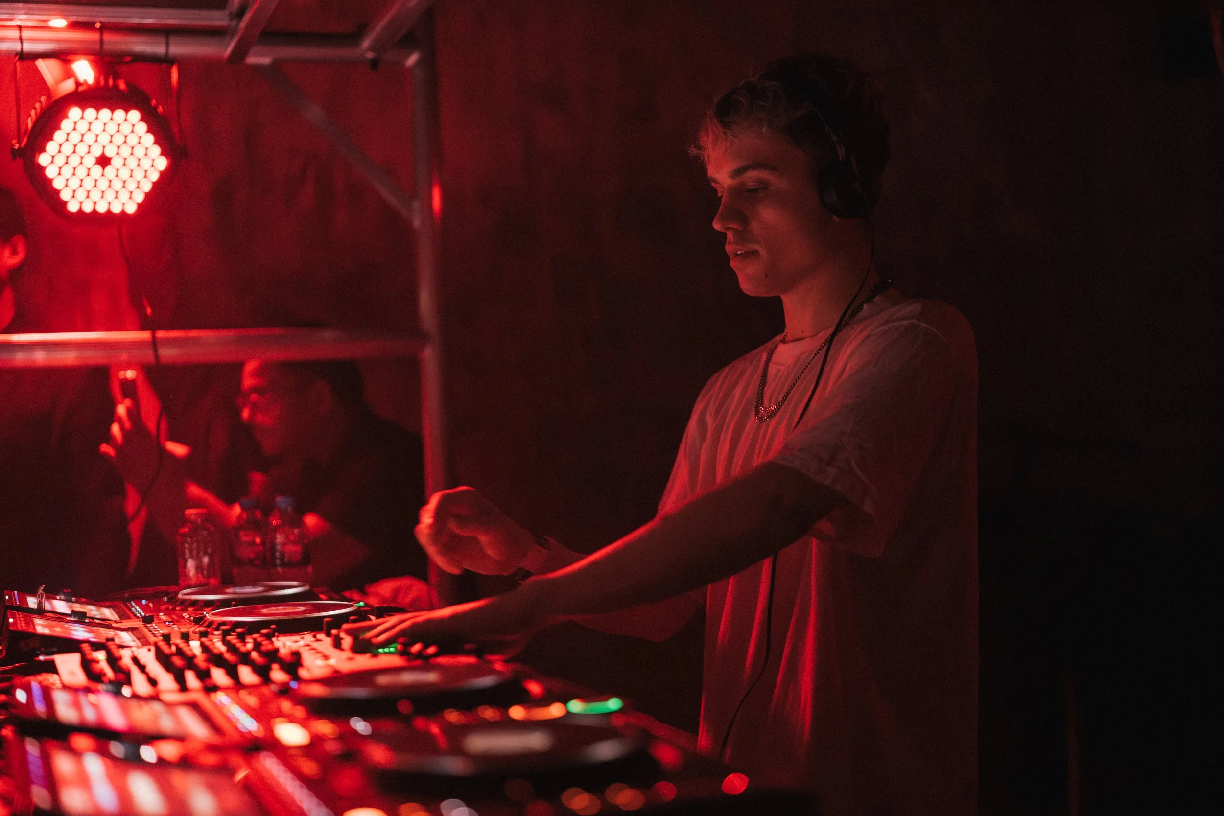 A young male DJ wearing headphones behind a mixing console at a dimly lit nightclub or party, with red lighting.