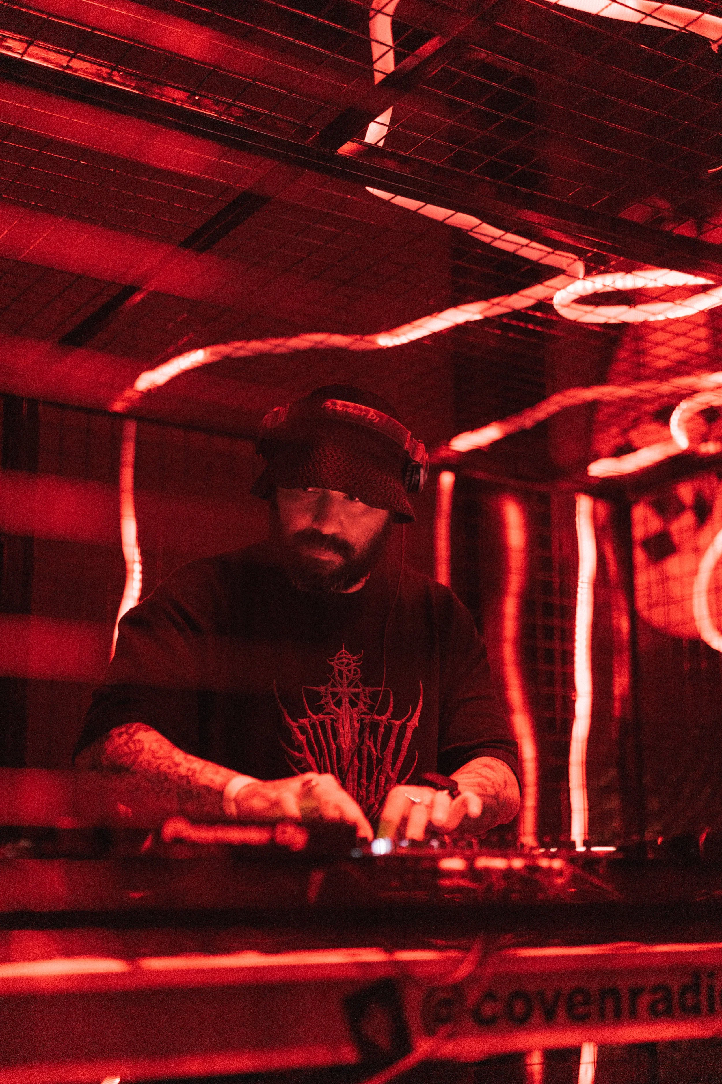A DJ wearing a black beanie, headphones, and a black shirt with red graphics, operating DJ equipment in a red-lit setting.