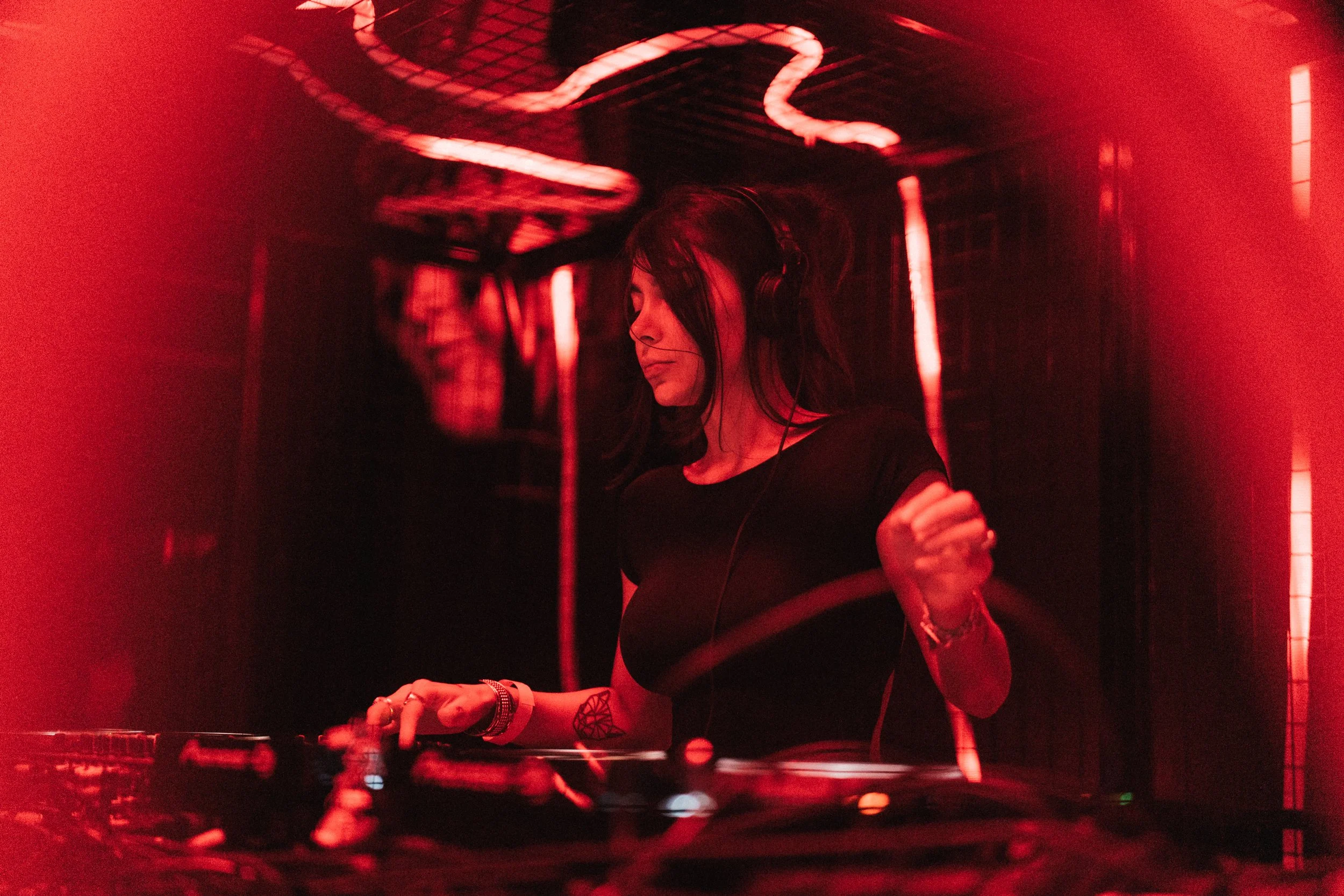 A female DJ with long dark hair and tattoos on her arms, wearing headphones, performing at a nightclub with red lighting and a neon snake-shaped light overhead.