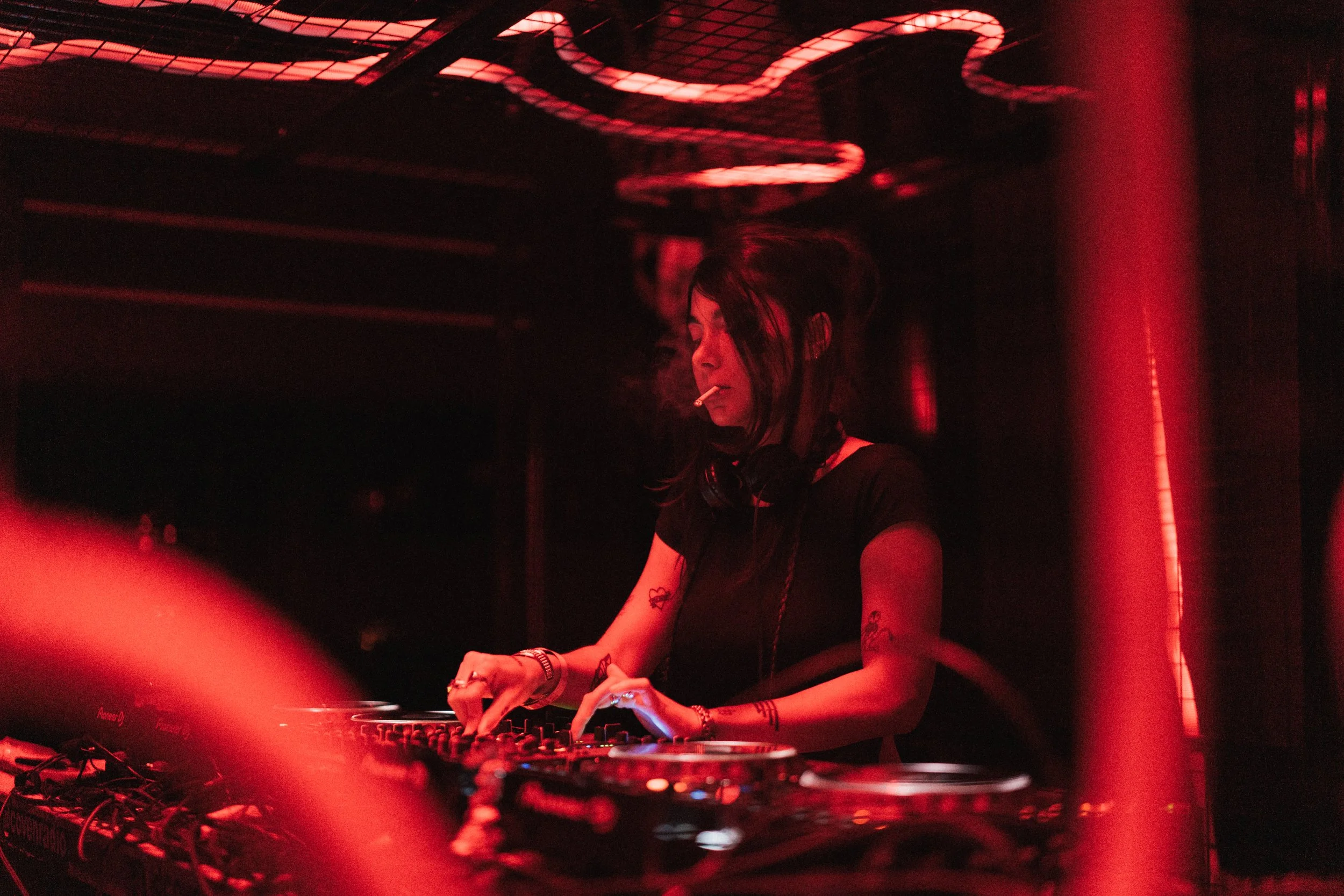 A female DJ with tattoos, wearing headphones around her neck and a black t-shirt, is working on her DJ equipment in a dark, red-lit environment. She is smoking a cigarette.