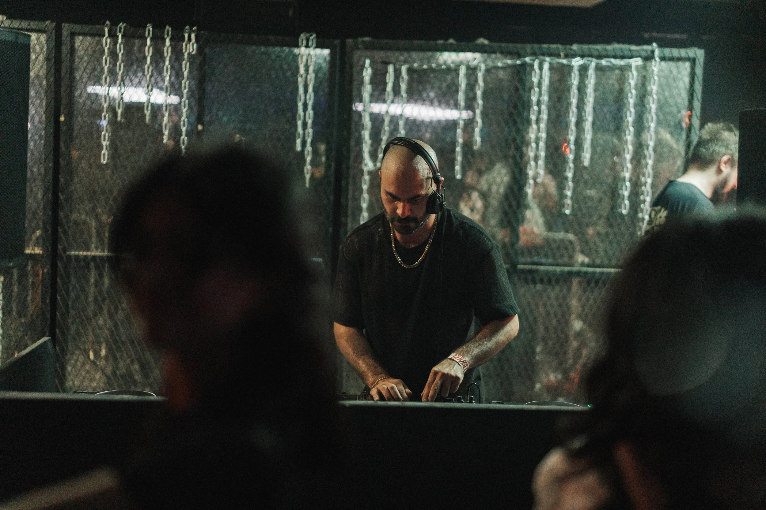 A DJ with a shaved head wearing headphones and a black t-shirt, standing behind DJ equipment in a dimly lit nightclub.