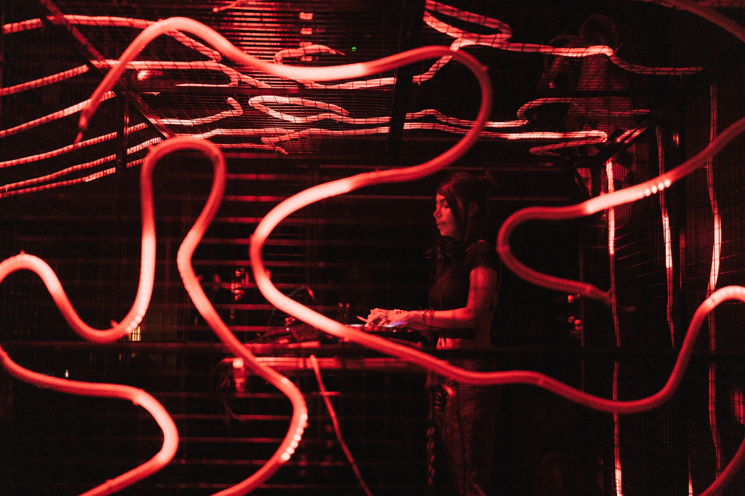 A woman with dark hair standing inside a dark room illuminated by red neon lights, with visible neon tubes and grid structures around her.