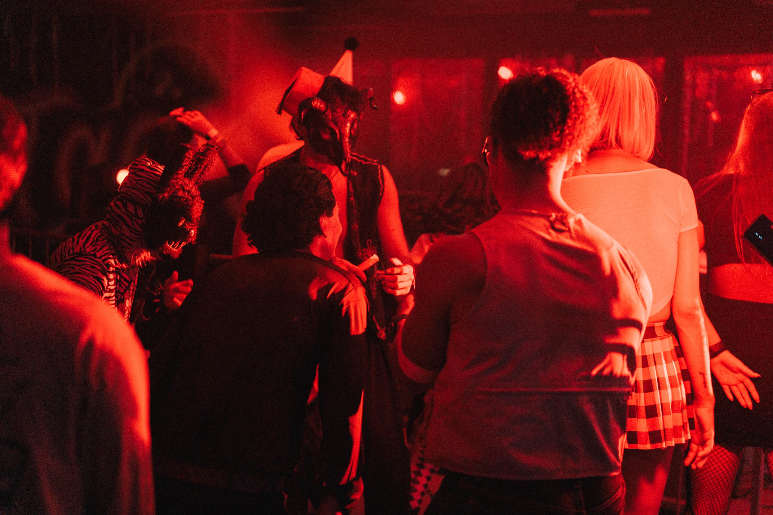 People dancing and socializing in a dimly lit club with red lighting, some wearing costumes and masks.