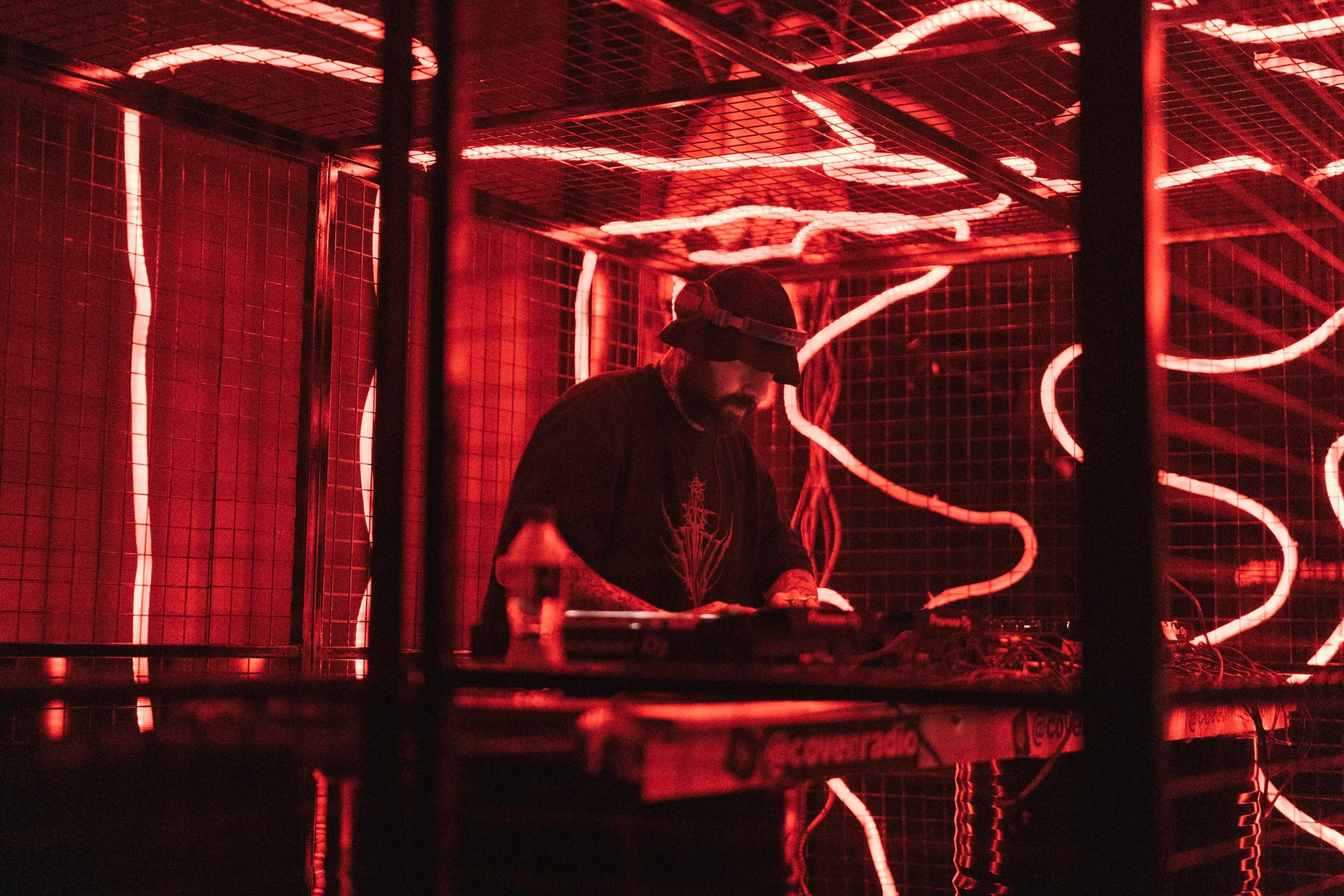 A DJ wearing a black cap and shirt with a graphic design, performing at a red-lit venue with neon lights and a grid-patterned background.