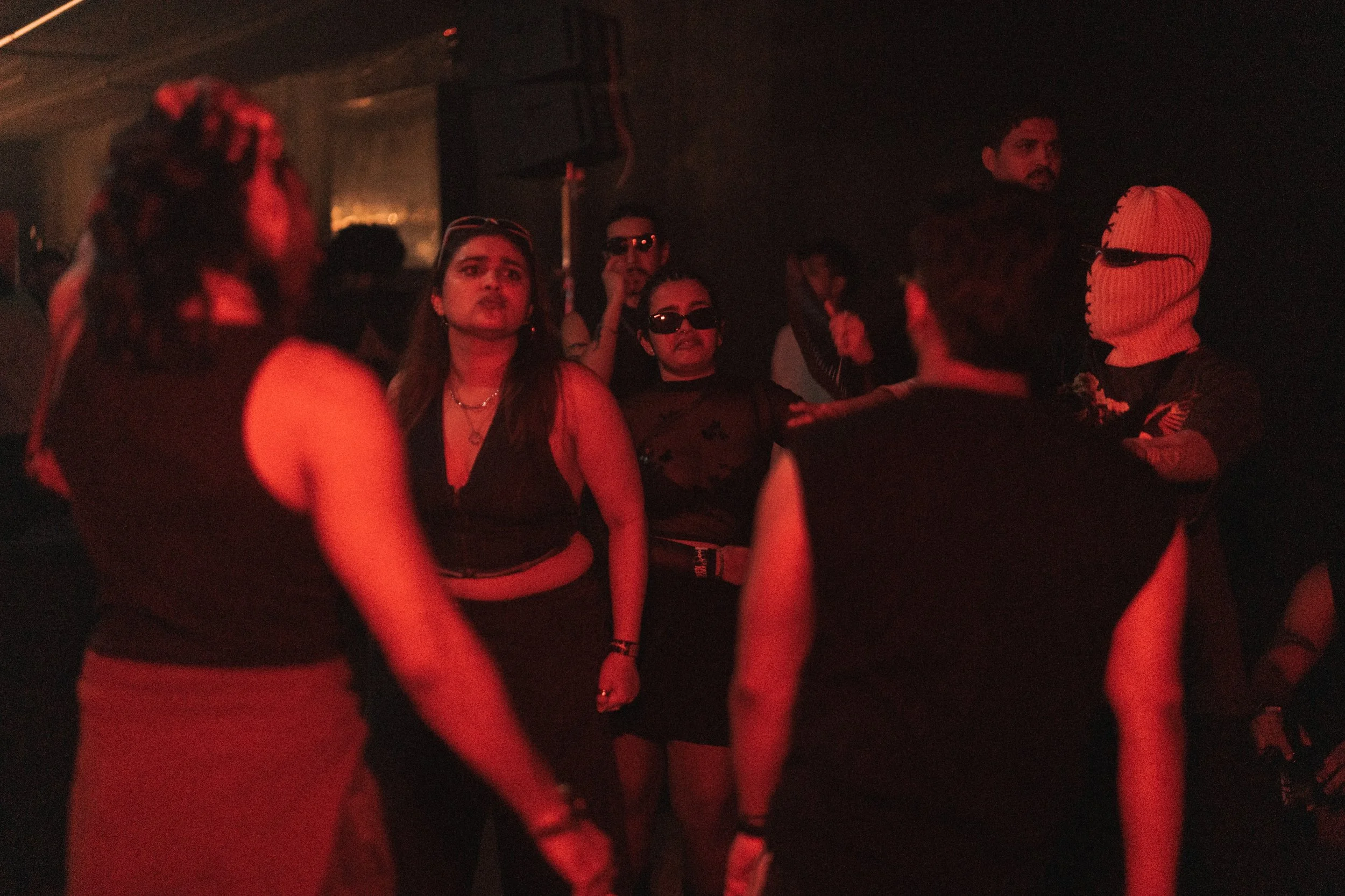 People gathered at a dark party or nightclub with red lighting, engaged in conversation or dancing.