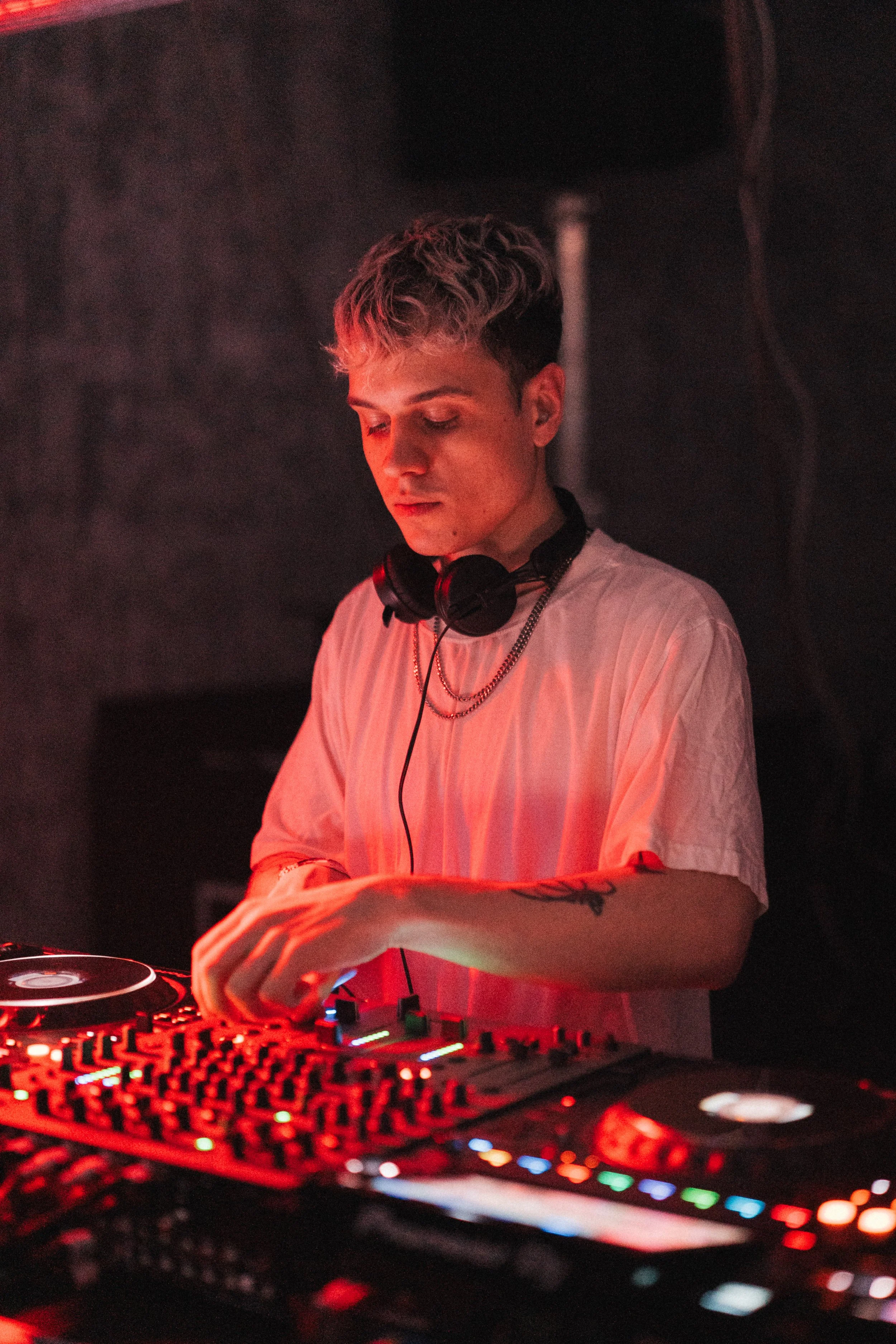 A DJ wearing a white shirt with headphones around his neck, concentrating on mixing music at a DJ controller with colorful lights.