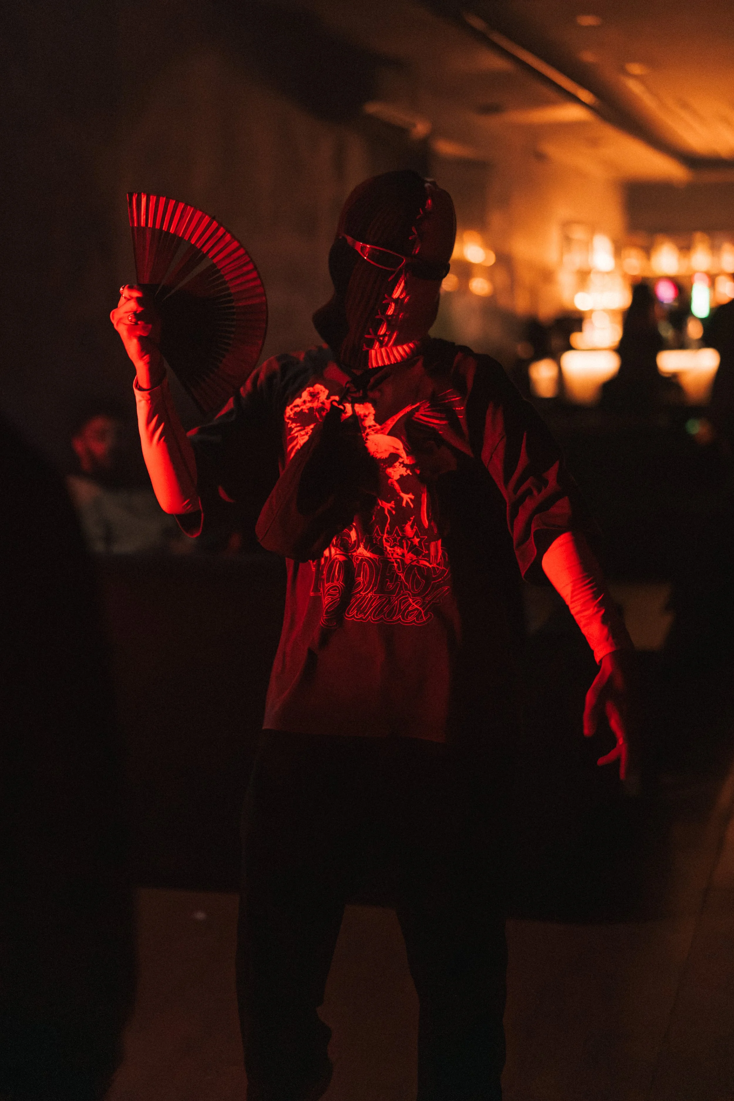 Person wearing a black mask with red lighting, holding a fan, in a dark indoor setting, possibly a nightclub or performance space.