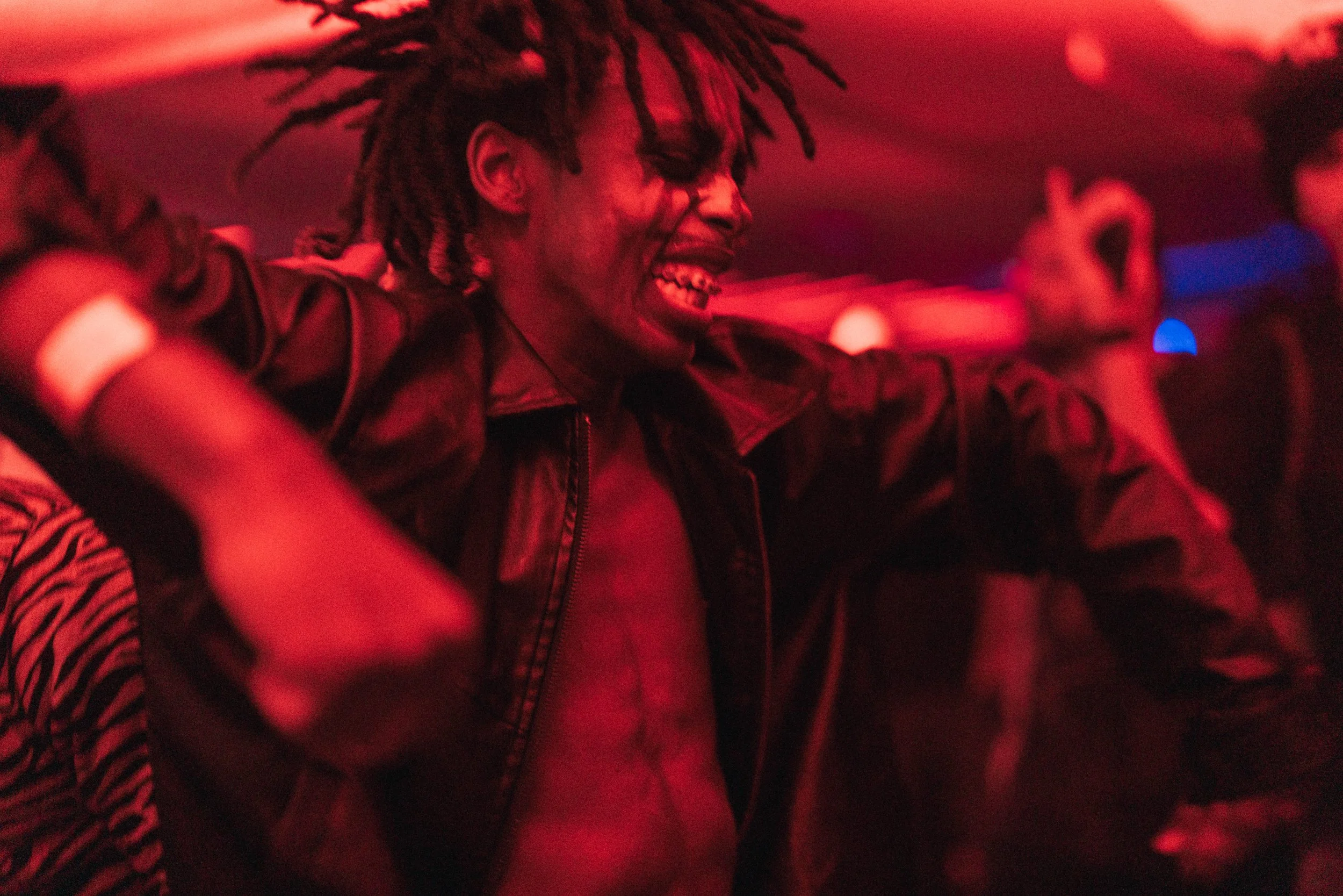 Person with dreadlocks, wearing an open leather jacket, smiling, in a dimly lit room with red lighting.