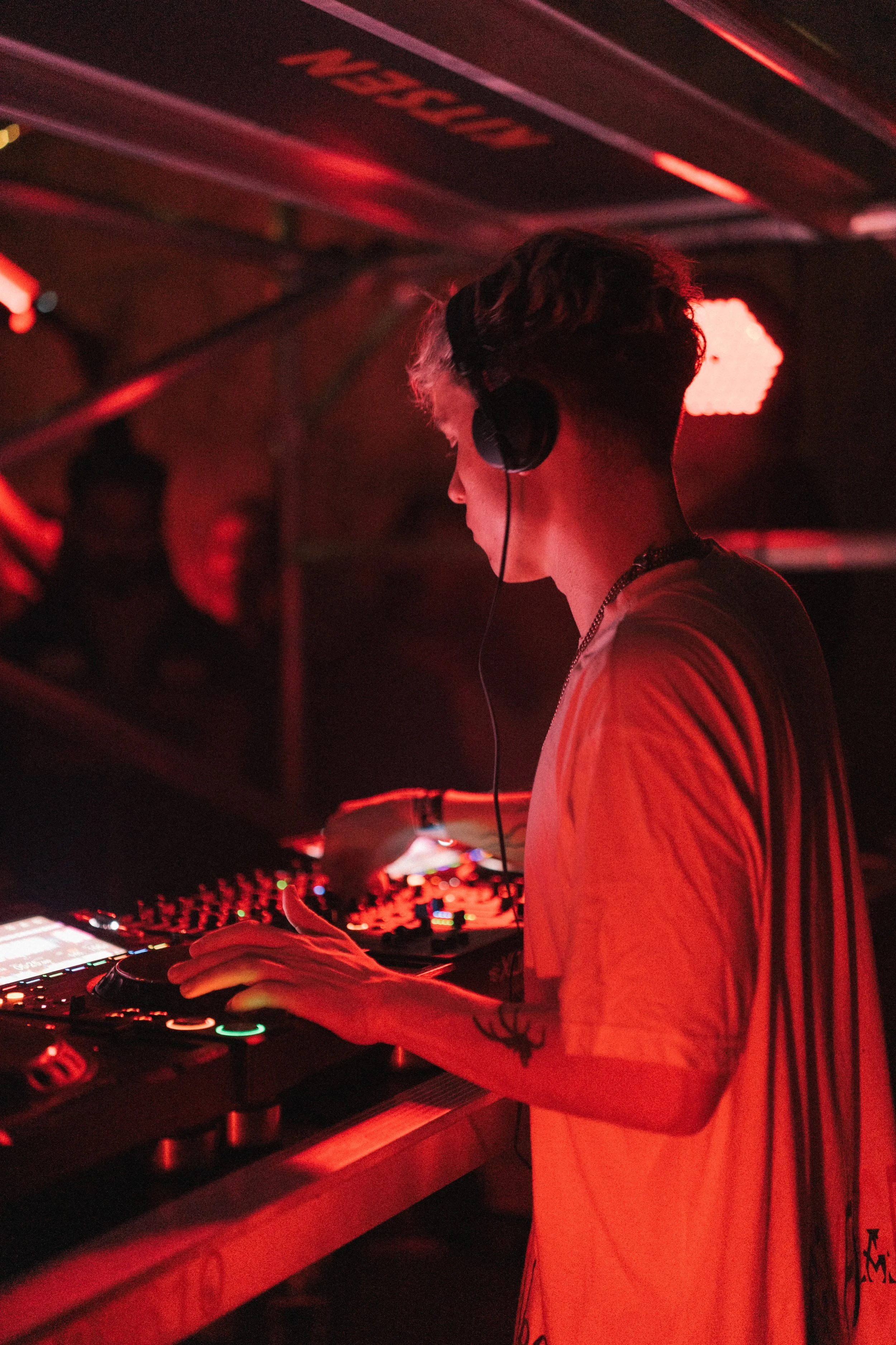 A DJ in a dimly lit club, wearing headphones and a striped T-shirt, is mixing music on a digital DJ controller, illuminated by red and orange stage lights.