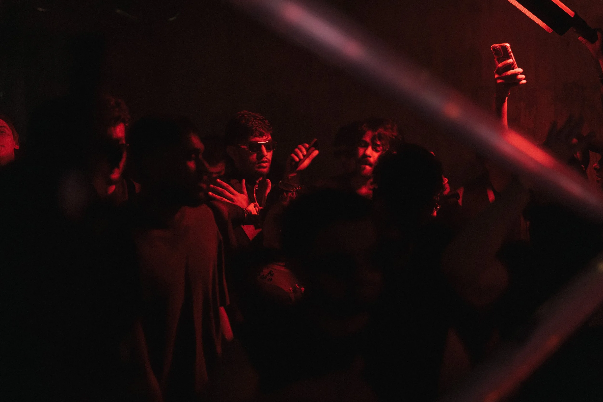 People dancing at a nightclub with red lighting, some wearing sunglasses and holding up a smartphone.