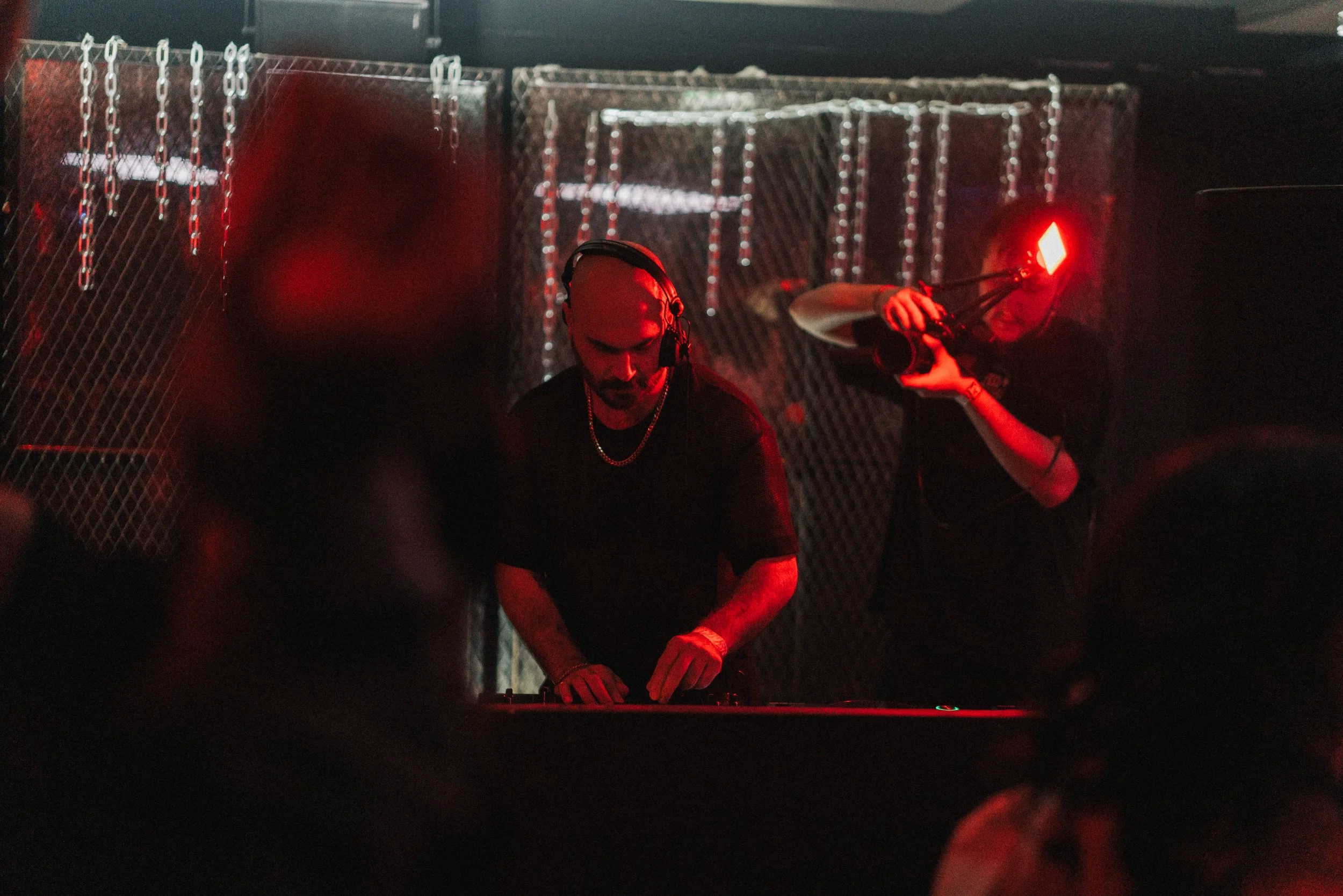 A DJ wearing headphones and a black shirt is performing, with a person behind holding a camera with a bright red light. The background consists of chain-link fencing with hanging light decorations, and the scene is illuminated with red lighting.