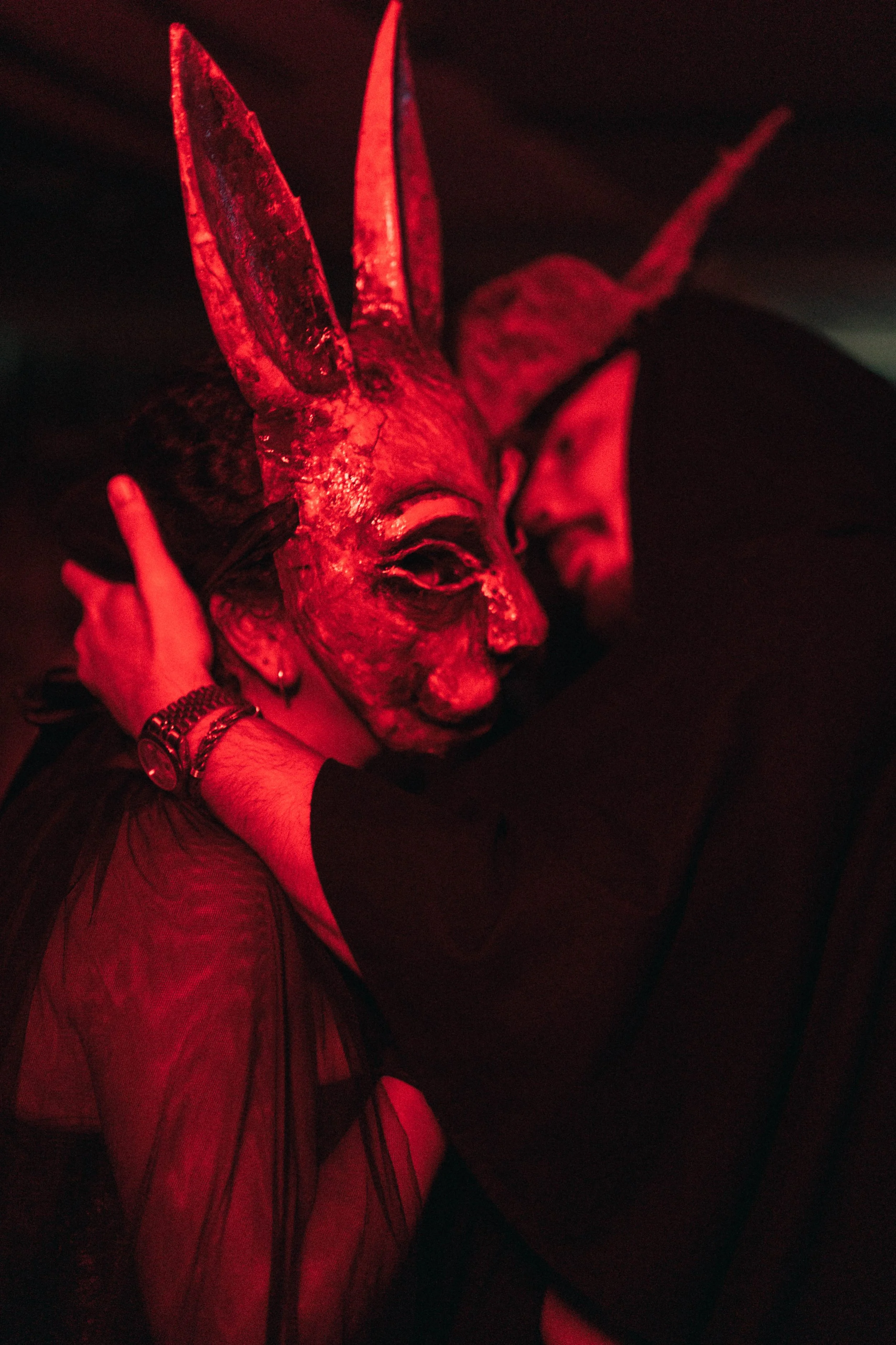 Person wearing a rabbit mask hugging another person in red lighting.