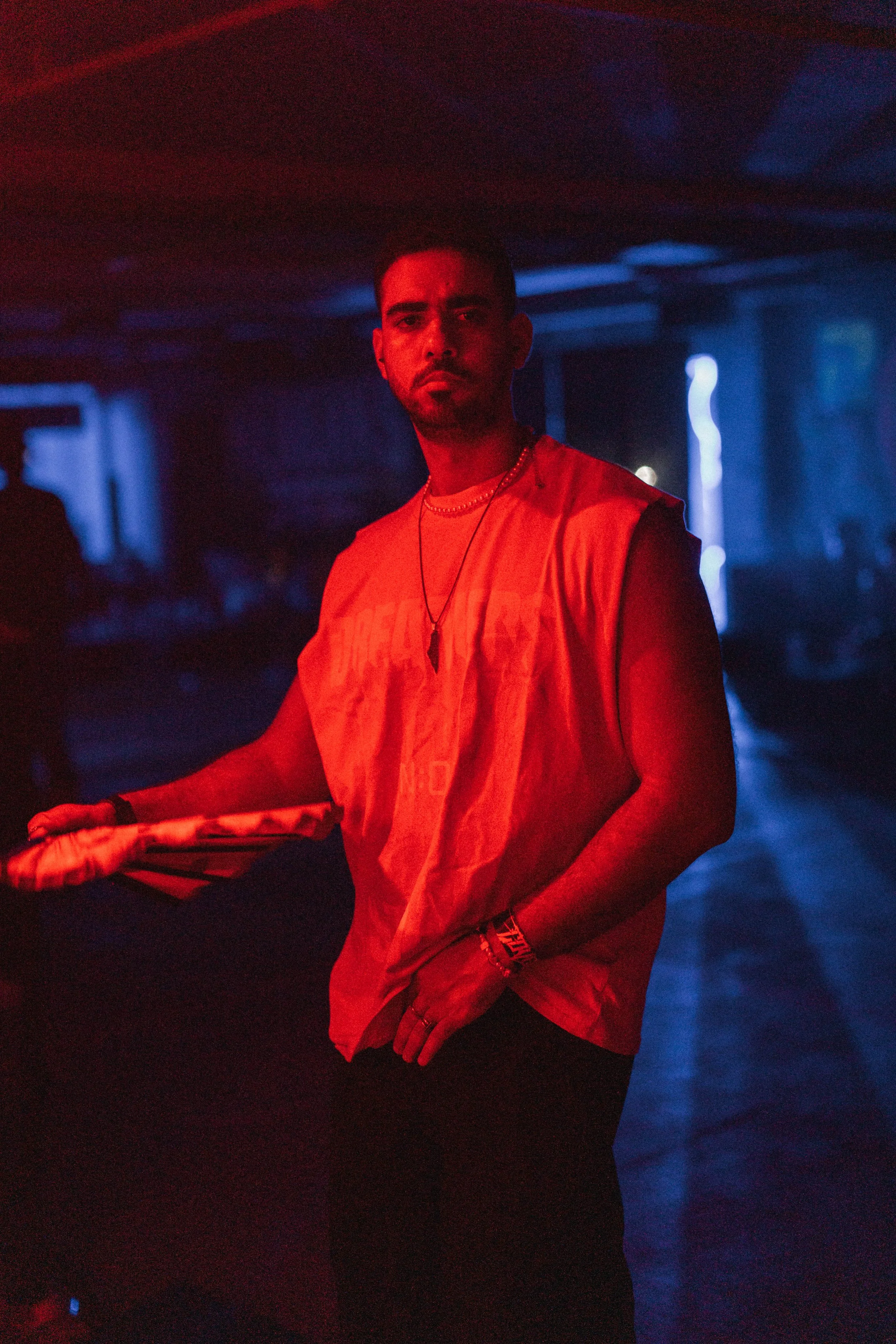 A man in a sleeveless orange shirt holding a tennis racket in a dark, colorful environment with red and blue lighting.
