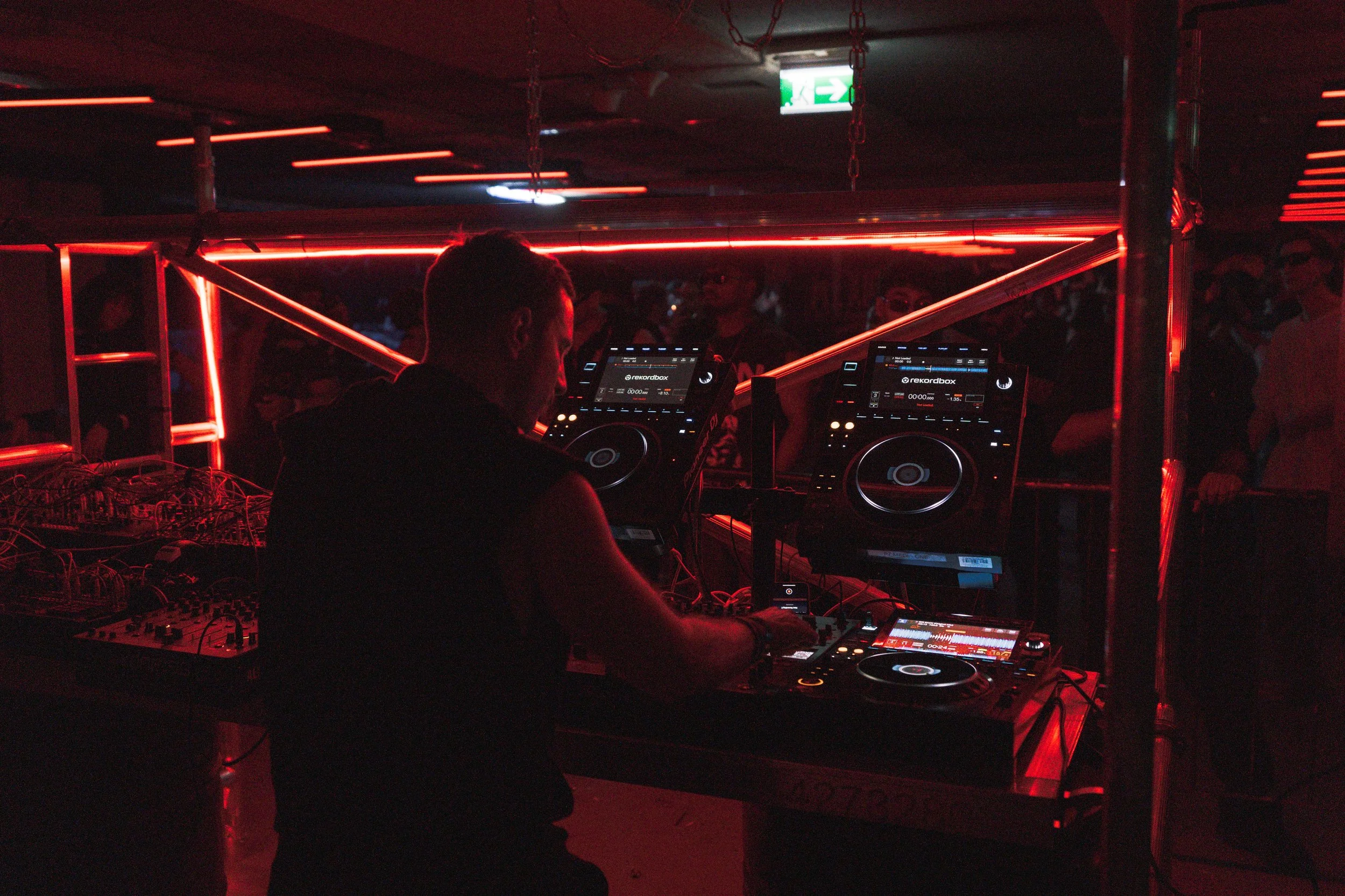 DJ performing at a nightclub with red neon lighting, surrounded by two large DJ decks and a crowd of people dancing in the background.
