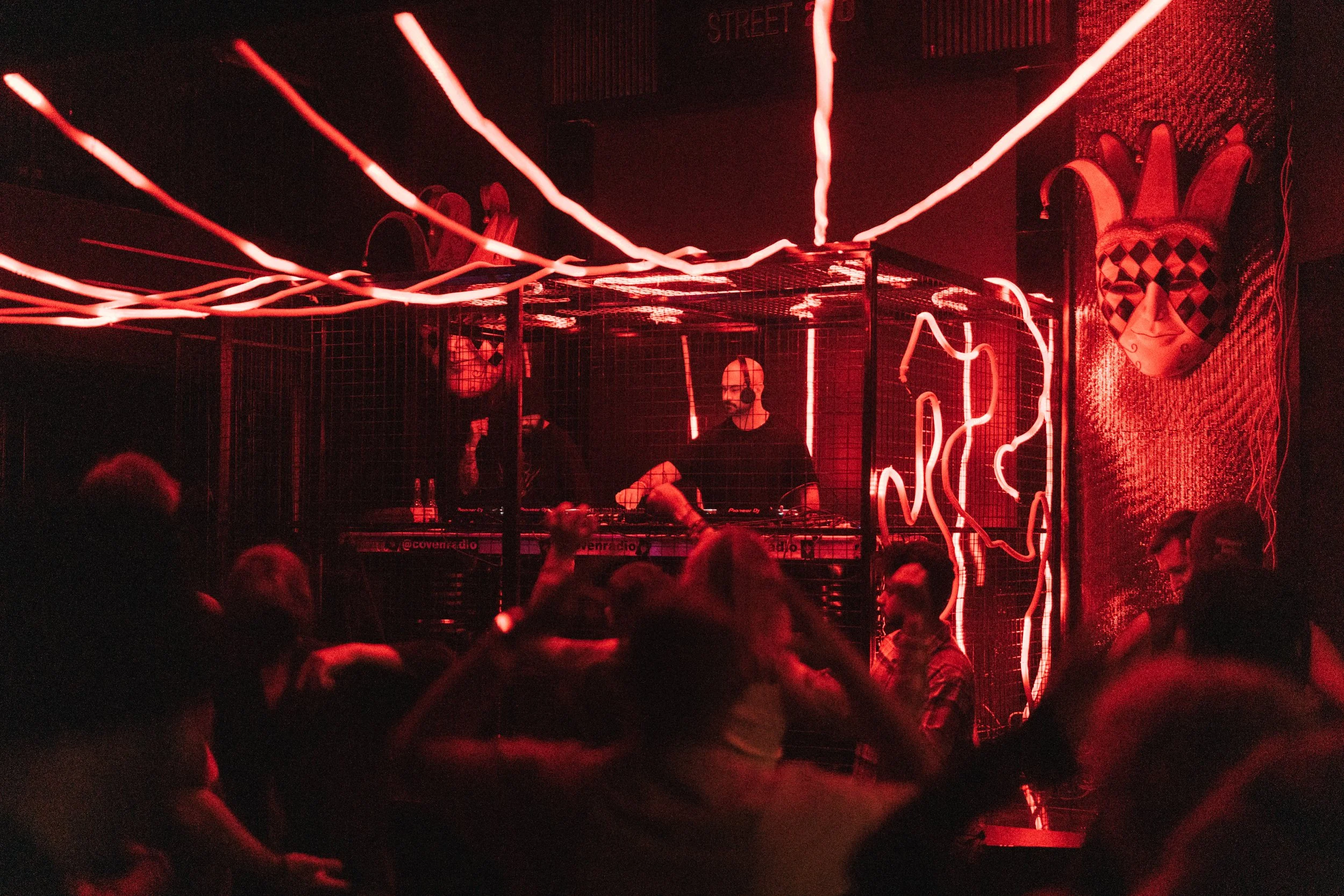 A DJ performs at a nightclub with red neon lighting, people dancing in front, and a mask sculpture on the wall.