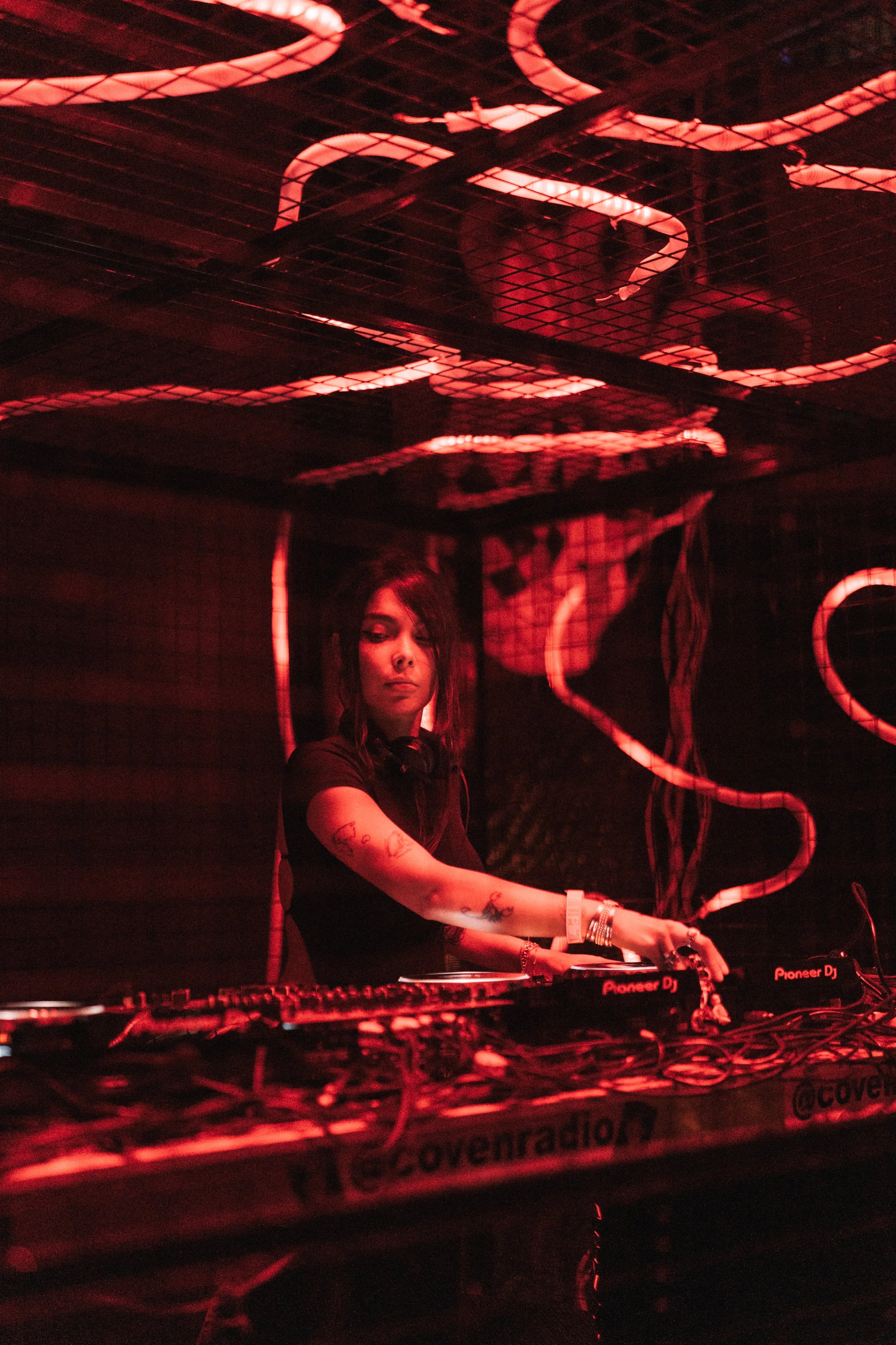 A female DJ with tattoos on her arms is performing at a nightclub, illuminated by red lighting with LED neon signs in the background.