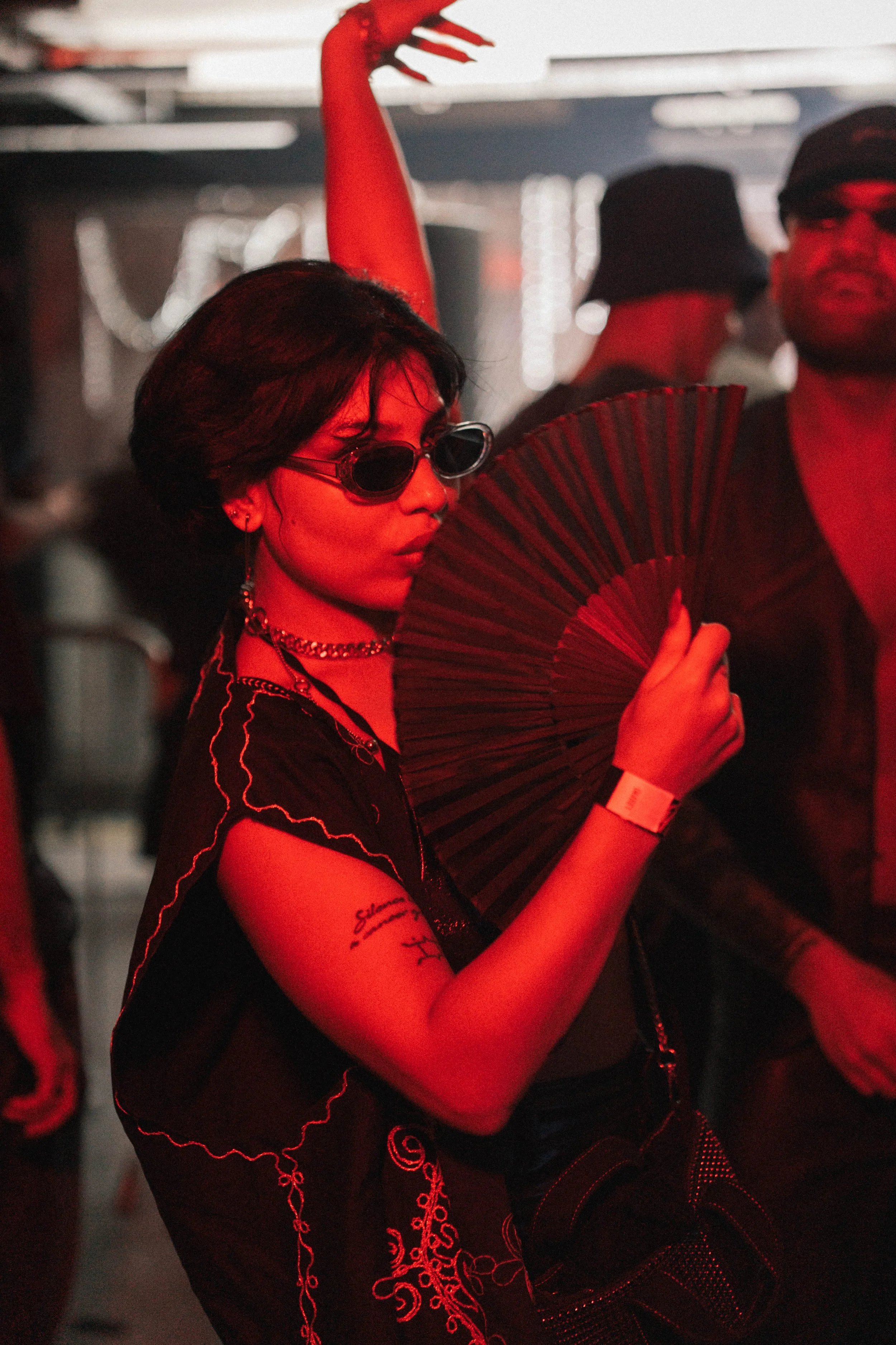 A woman with short dark hair, wearing sunglasses, earrings, and a black shirt with embroidered details, holds a black folding fan near her face. She is in a dimly lit environment with red lighting, accompanied by other people in the background.