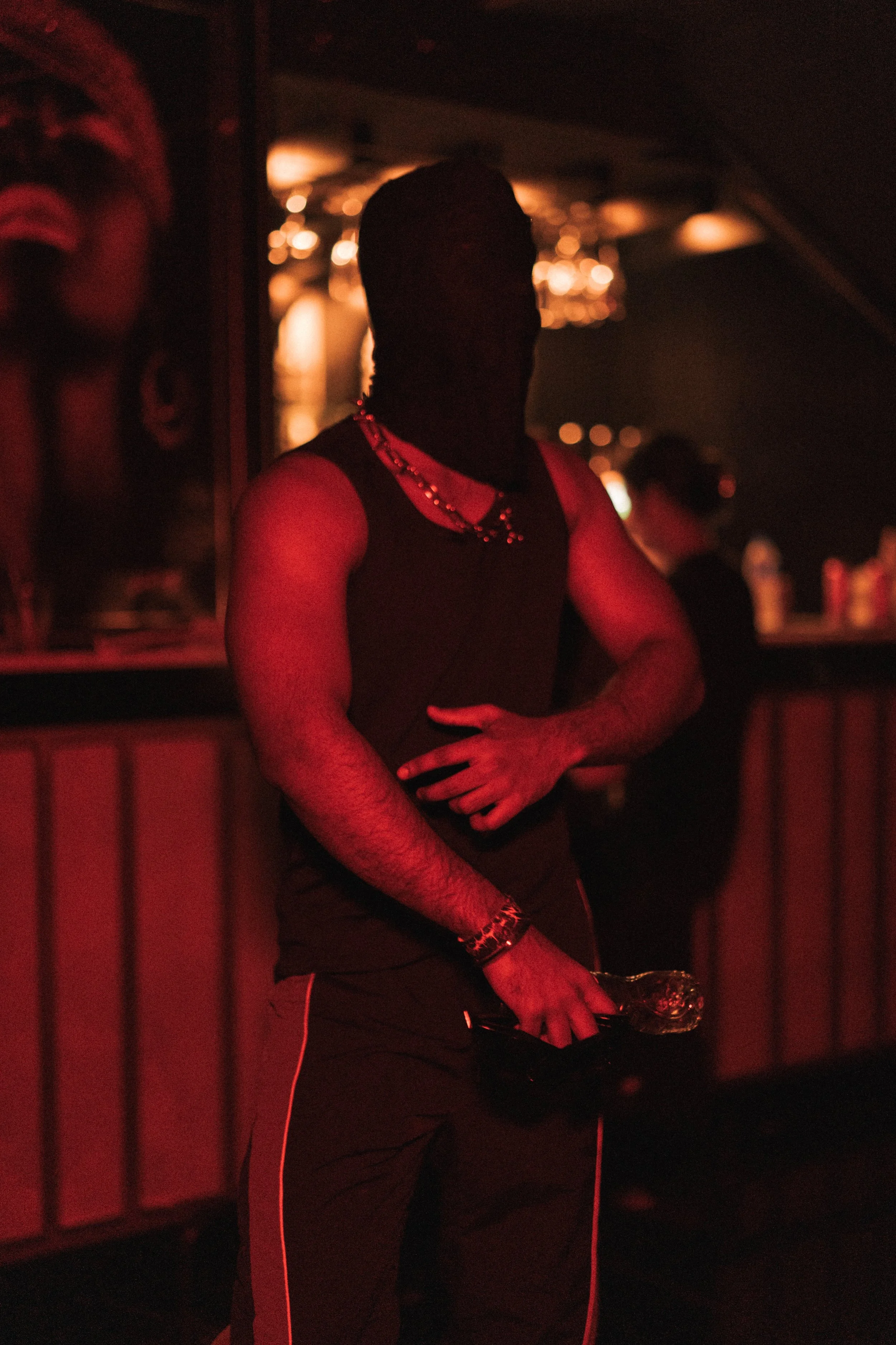 A man dressed in dark clothing with jewelry, holding a glass, in a dimly-lit bar with red lighting.
