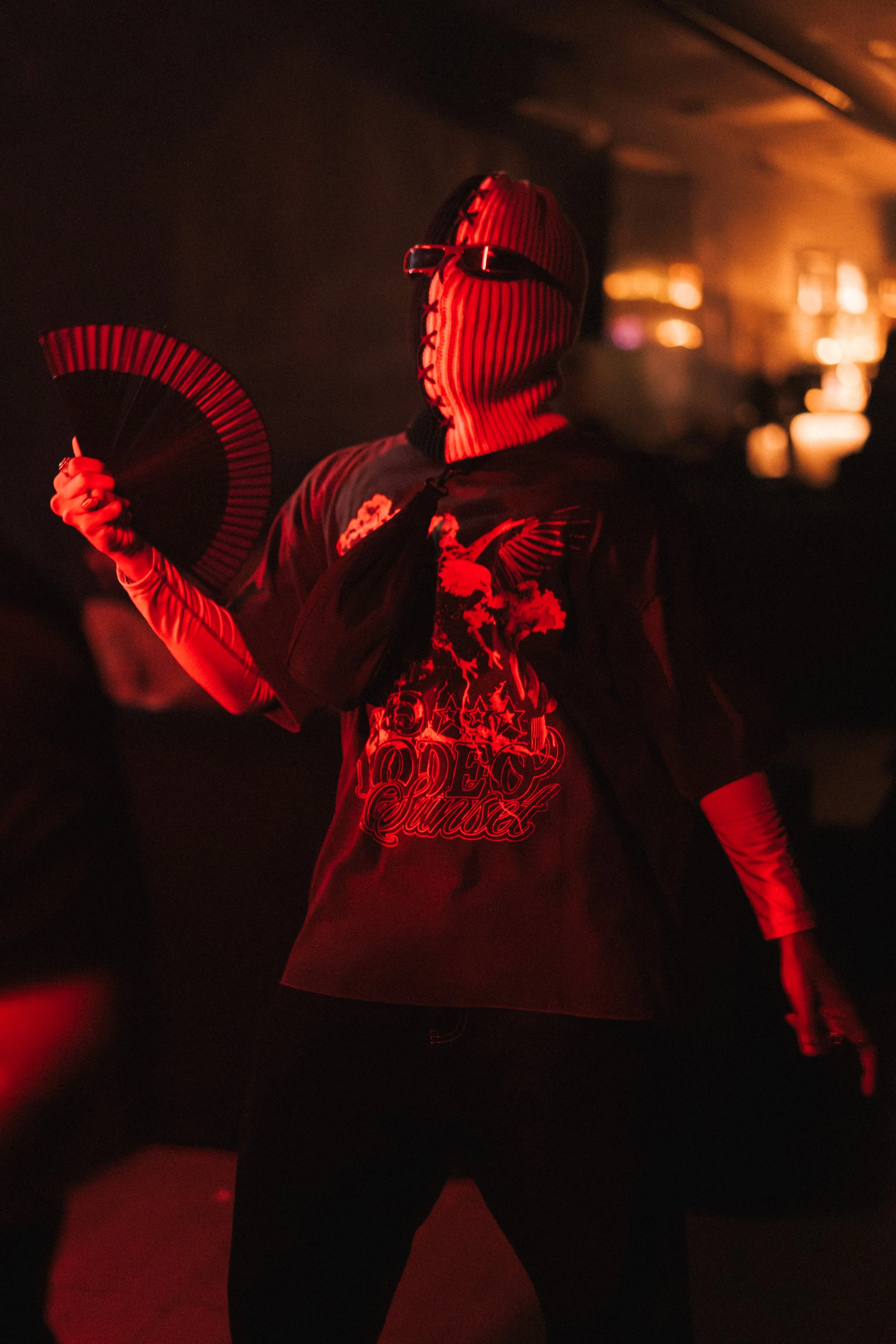Person wearing a red and black striped face mask with glasses, holding a fan, illuminated in red light, dark background, wearing a t-shirt with graphics and text, long gloves, in a dimly lit indoor setting.