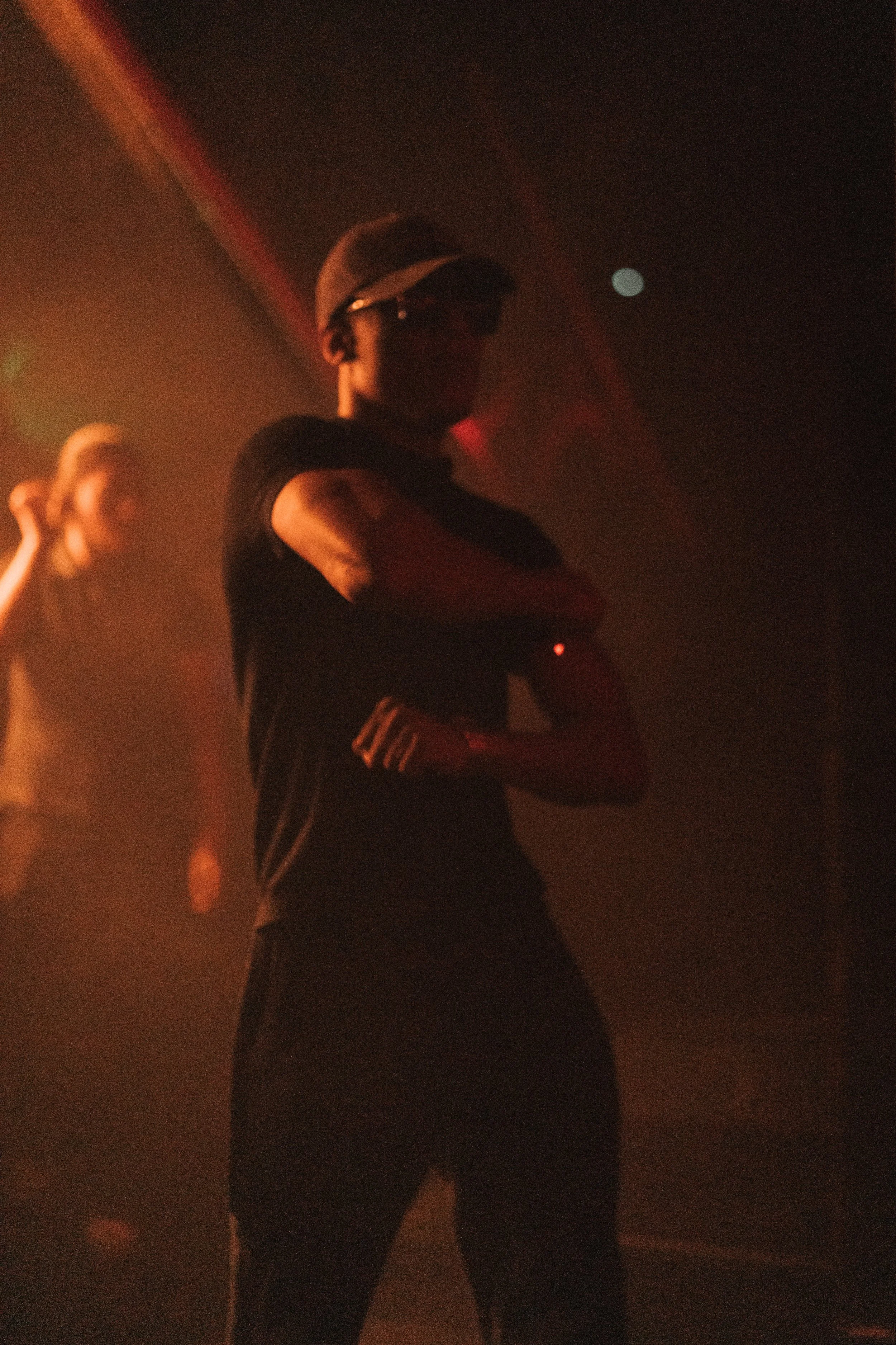 A person standing with arms crossed, wearing glasses and a cap, in a dark, smoky environment with a dim light.