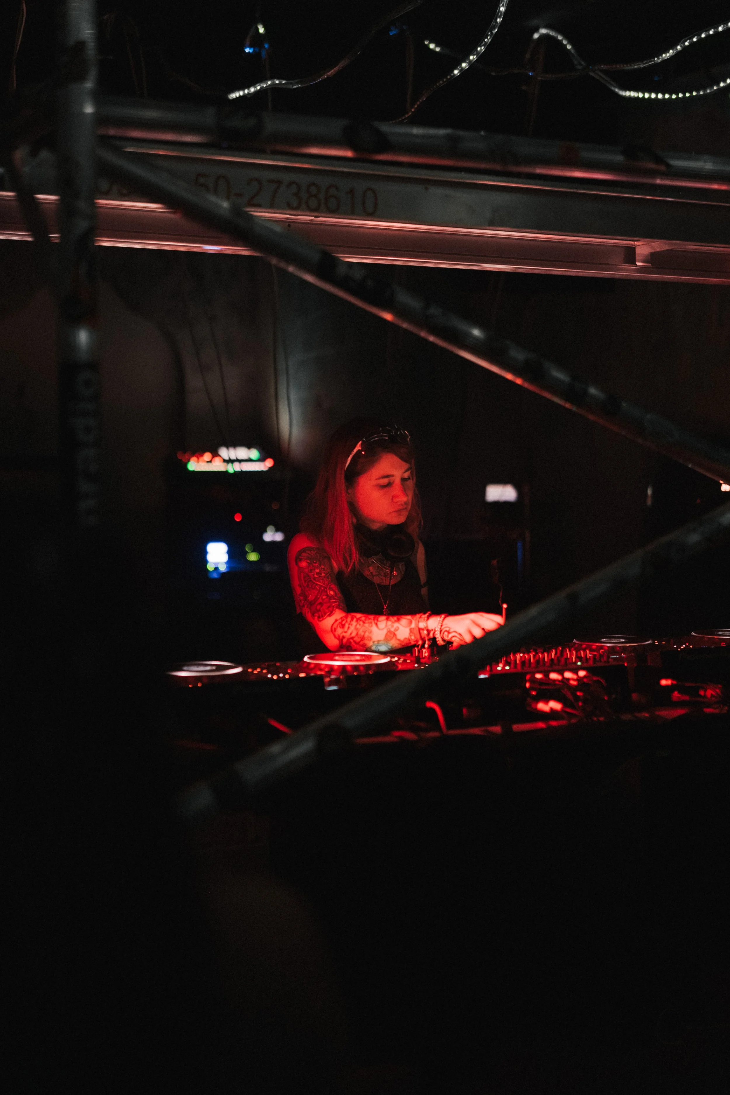 A female DJ with tattoos, wearing headphones around her neck, playing music at a dimly lit venue with red lighting.