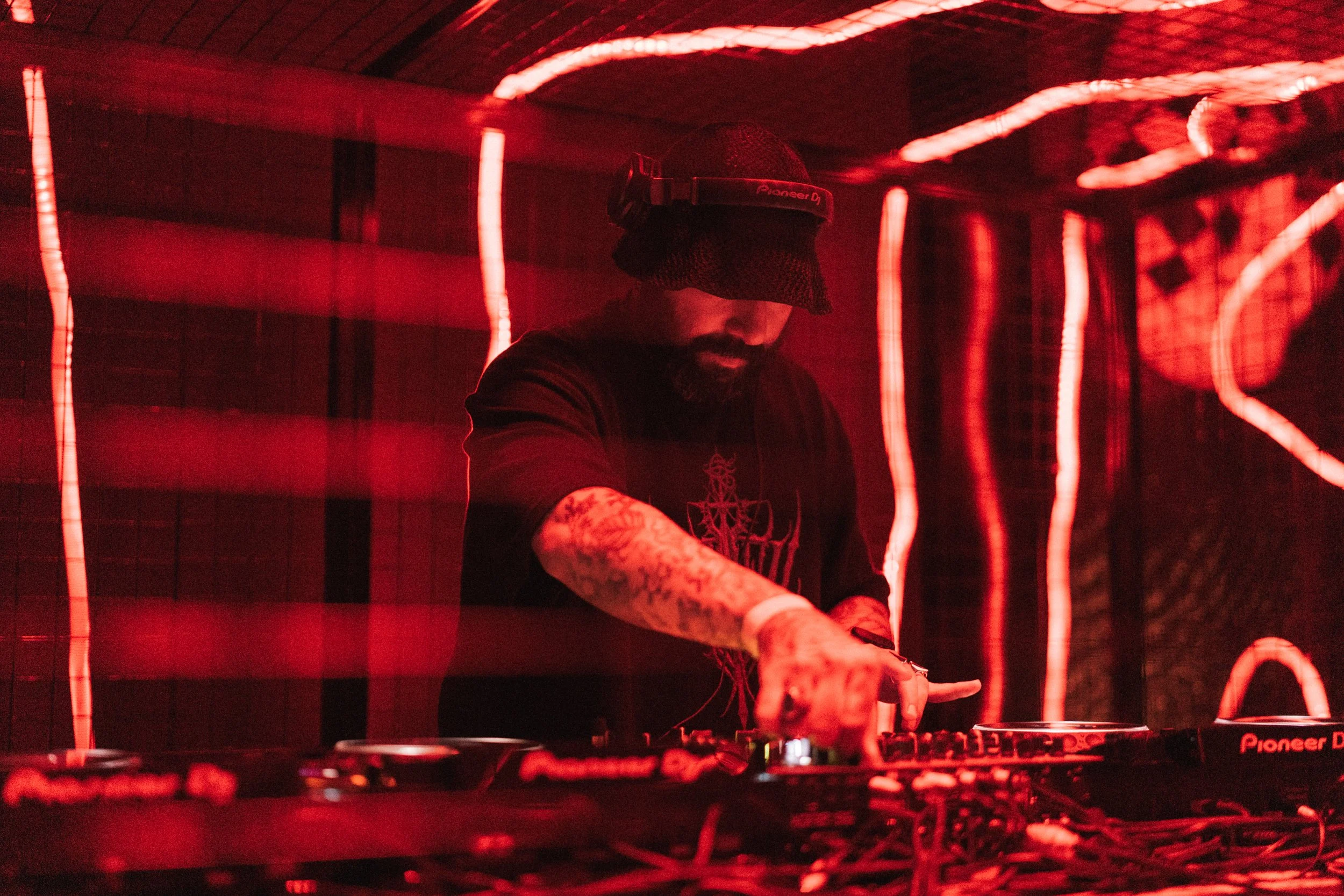 A DJ wearing a black hat and headphones is performing in a dimly lit space with red lighting and neon light strips on the wall behind him.