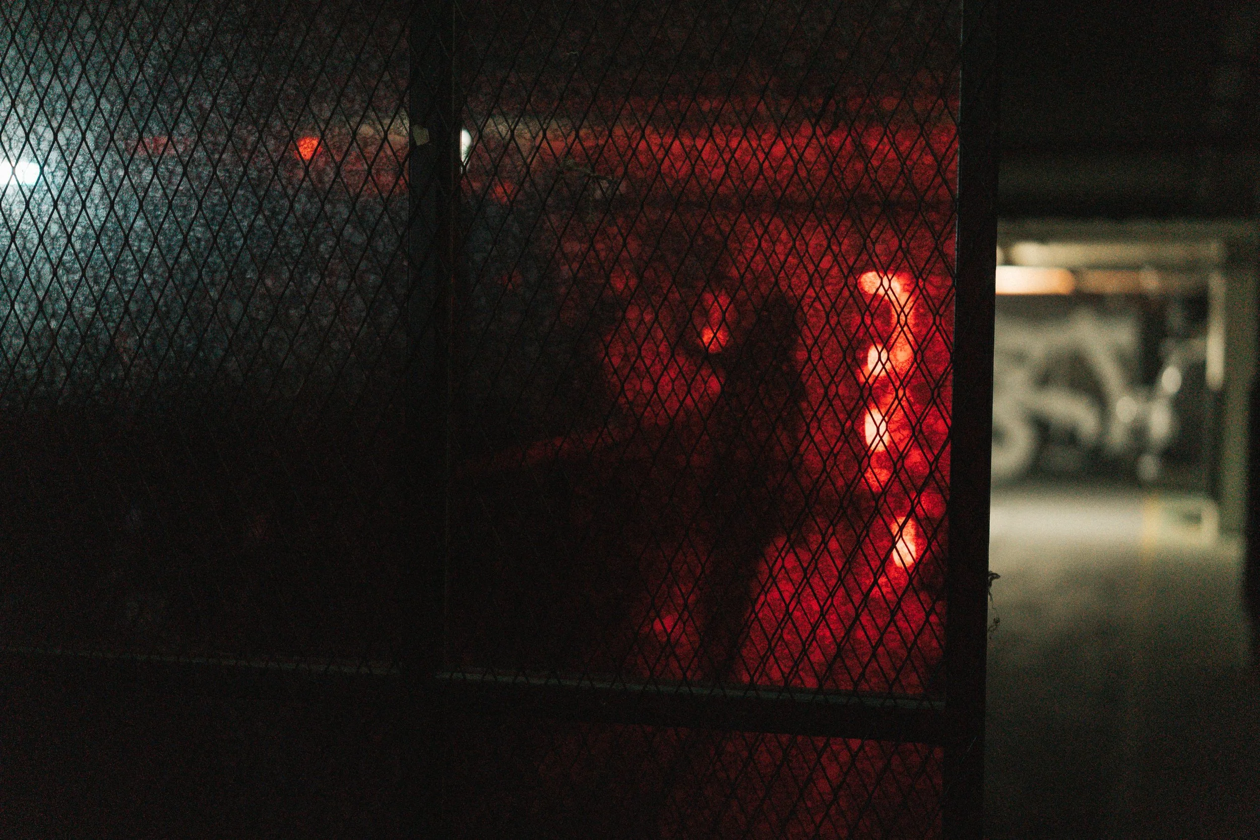 Dark parking garage viewed through a wire mesh fence with red and white lights in the background.