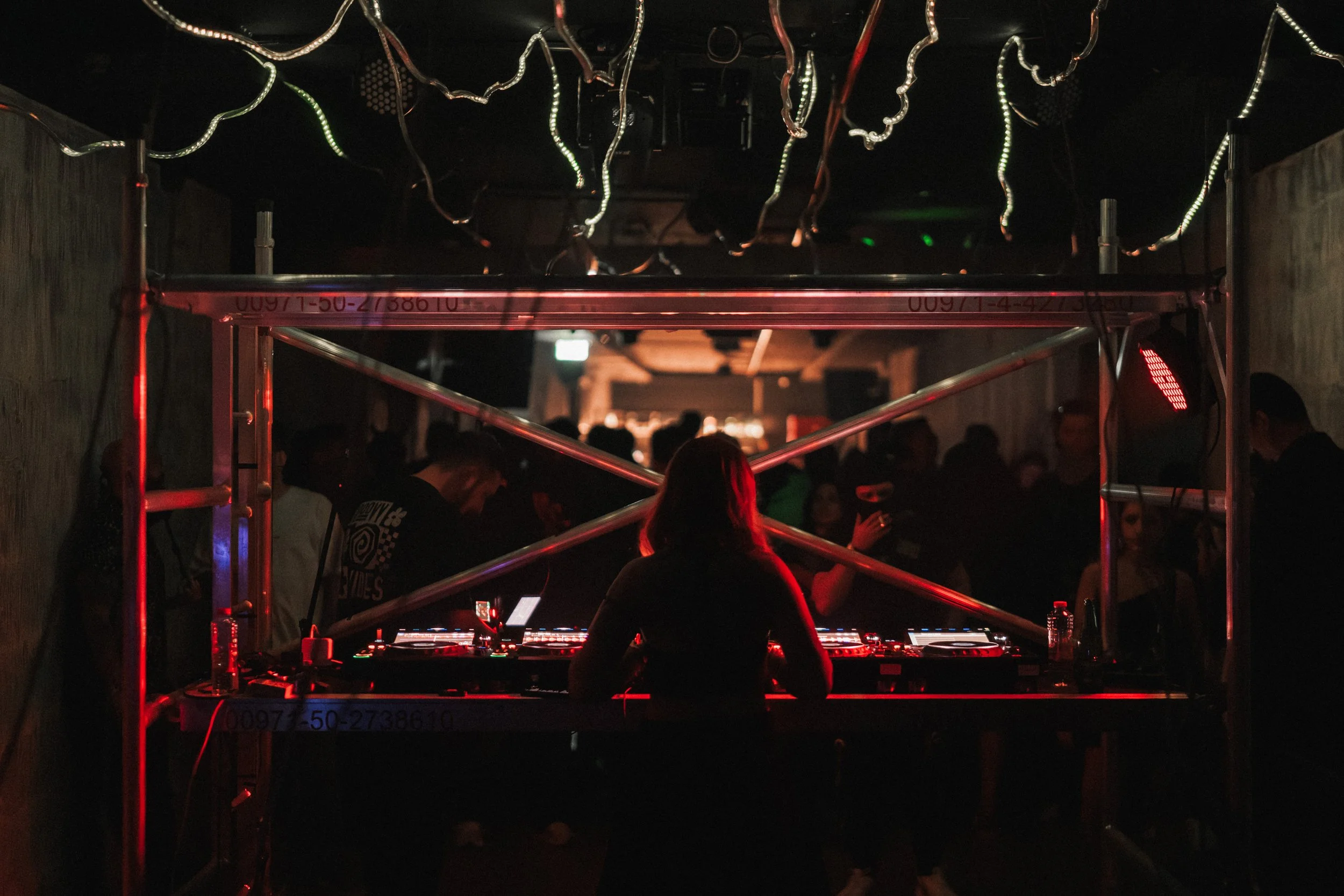 DJ performing at a nightclub with a crowd dancing in the background, dimly lit with red and green lights.