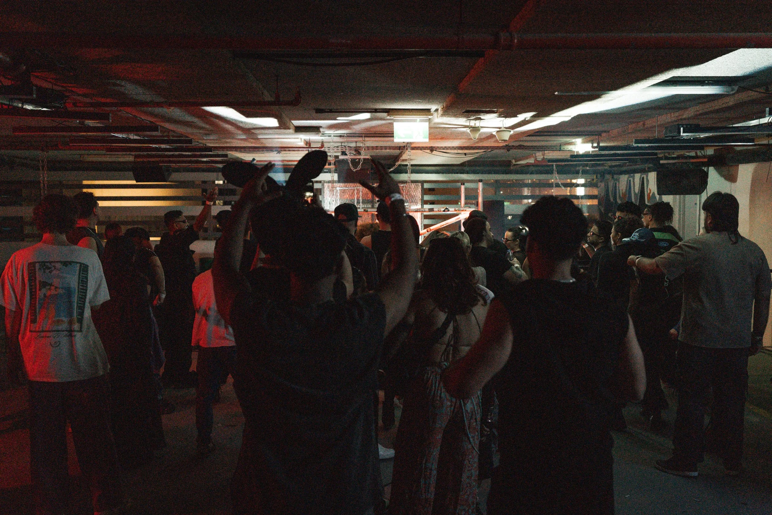 A crowded indoor concert or event venue with people facing the stage, including some with raised hands, under dark lighting with a lit stage at the front.