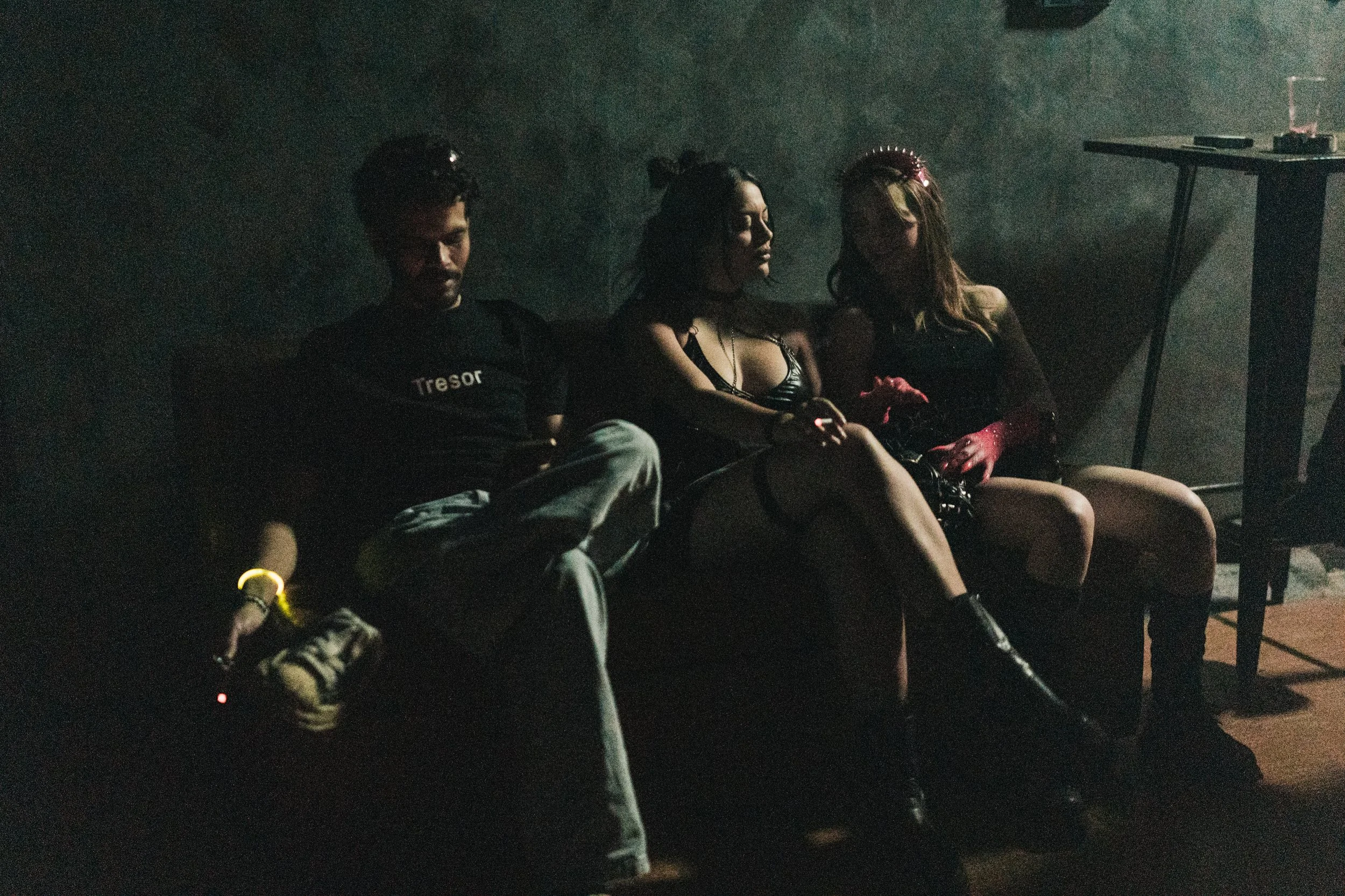 Three young adults sitting on a dark, dimly lit couch in a grunge-style room with no visible background details.