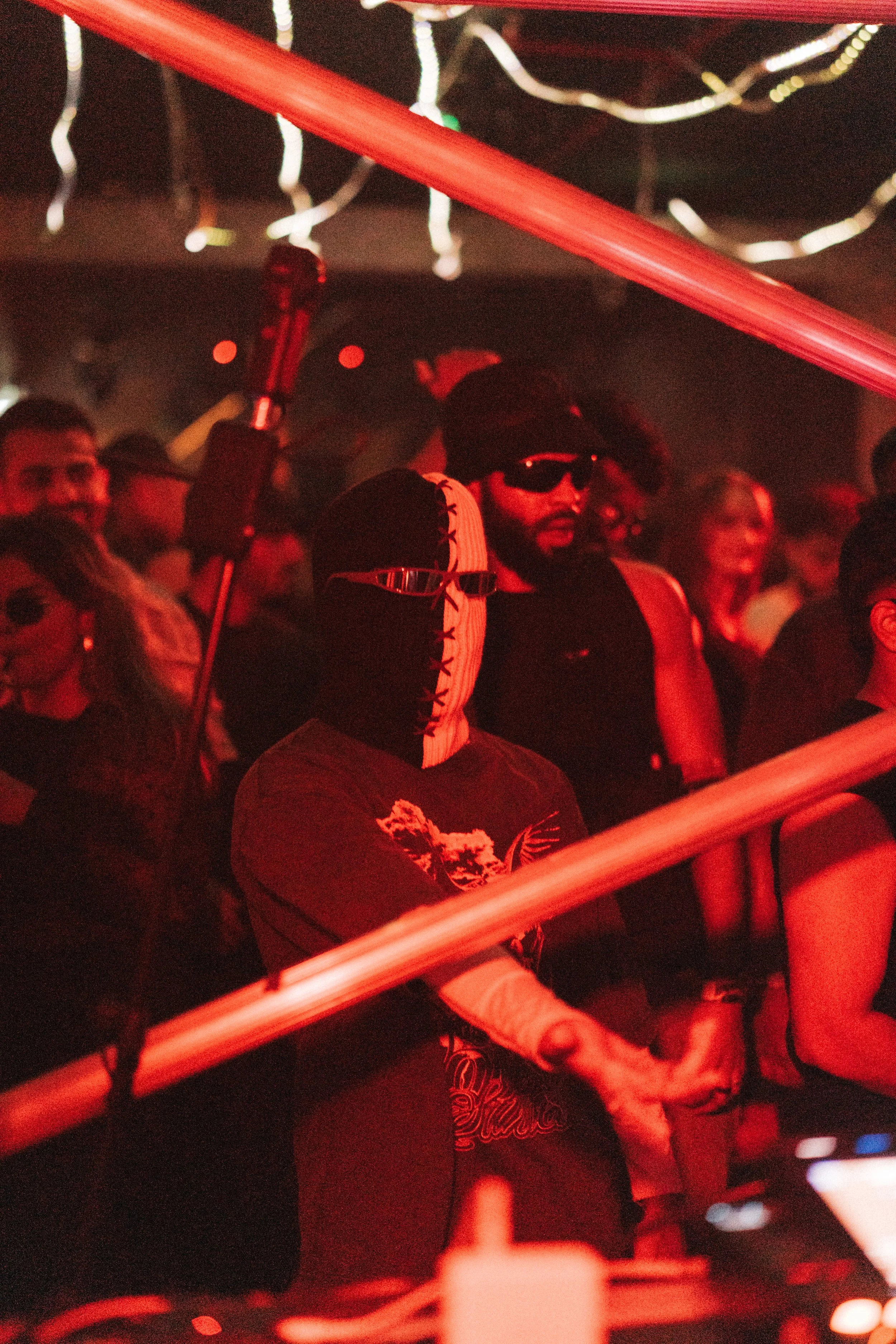 A person wearing a black and white mask, sunglasses, and a black hoodie stands among a crowd at a concert or event, illuminated by red lighting.