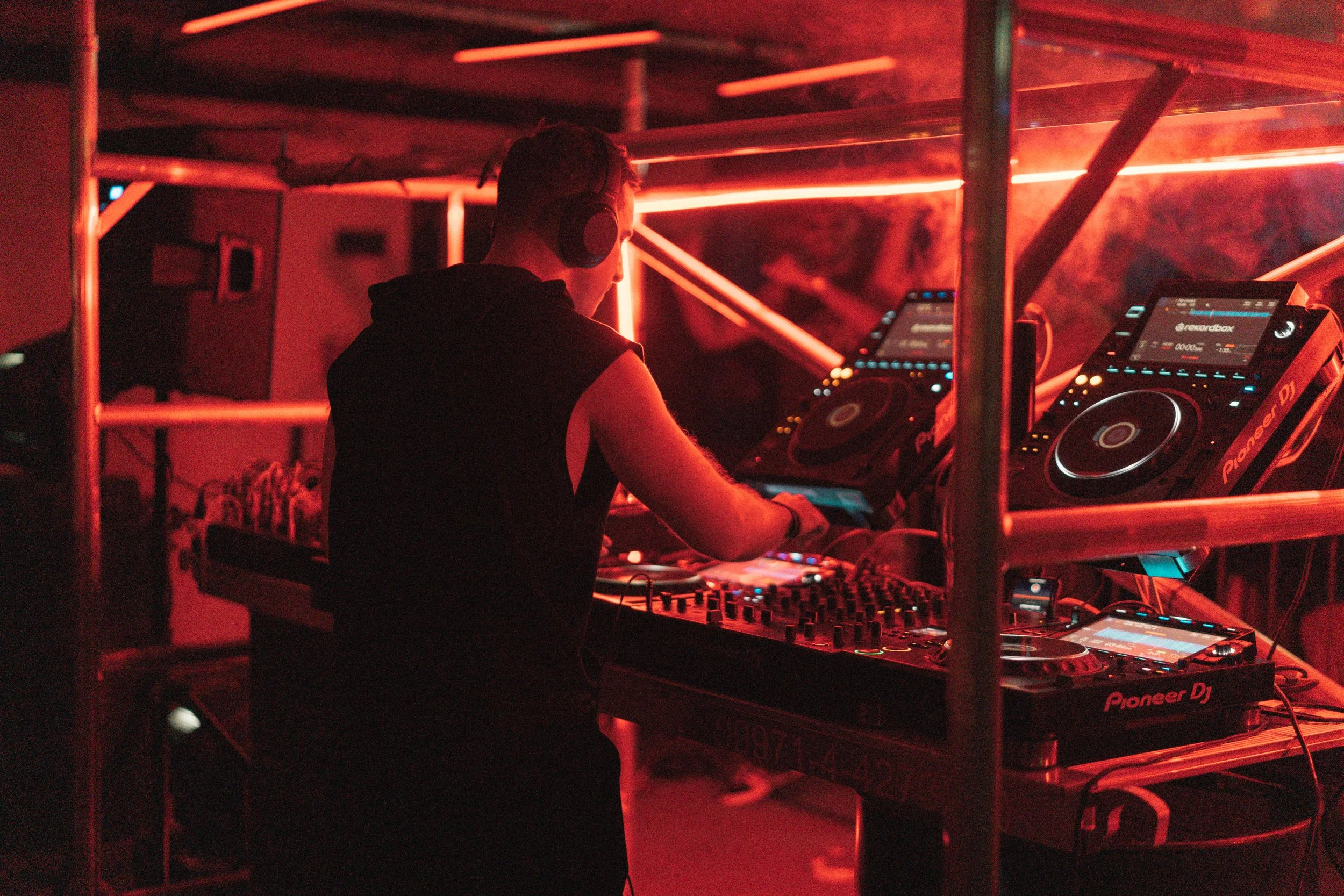 A DJ wearing headphones is performing at a club with red lighting, surrounded by DJ equipment including Pioneer CDJs and a mixer.