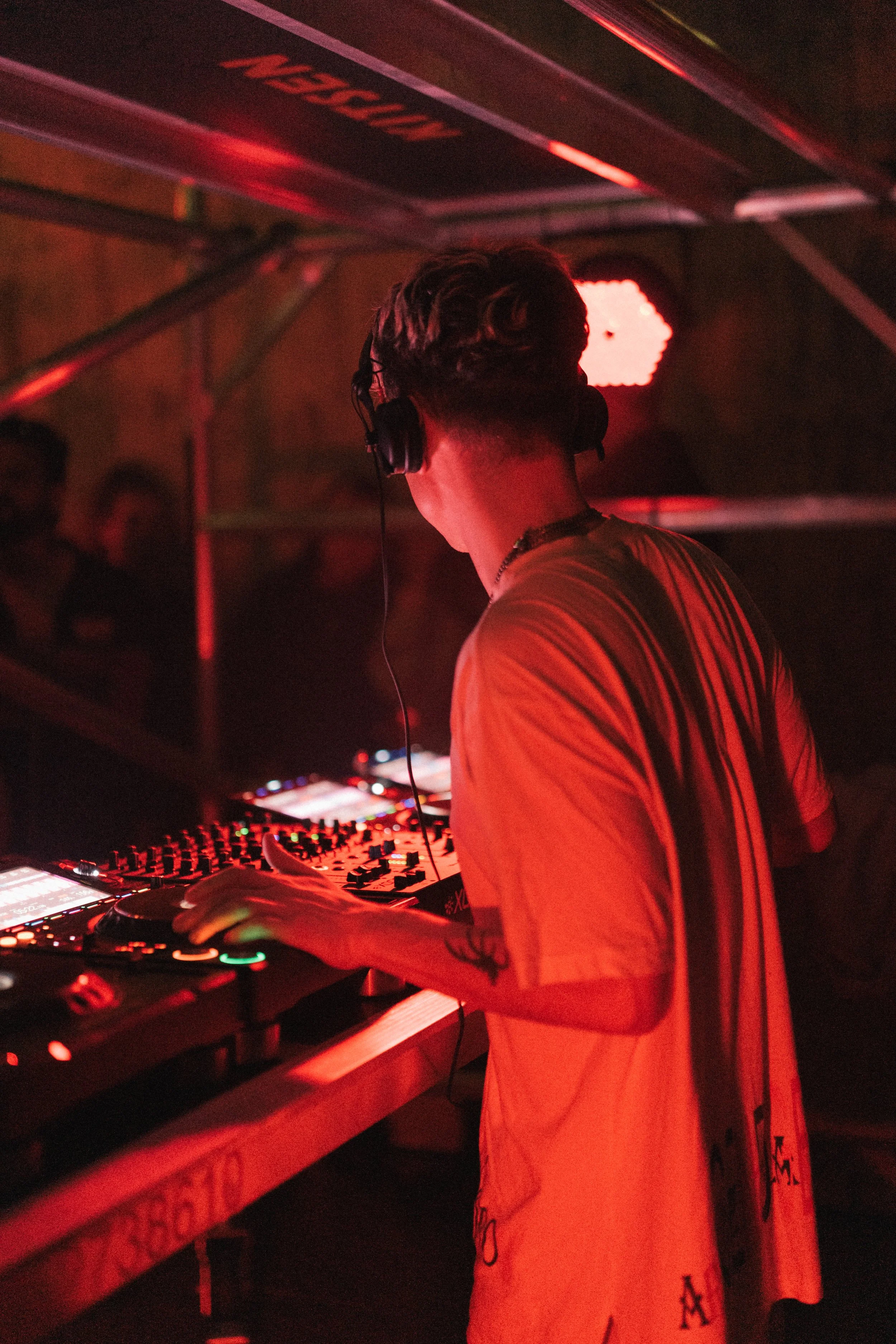 A DJ with headphones stands at a mixing console in a dimly lit club, illuminated by red lighting.