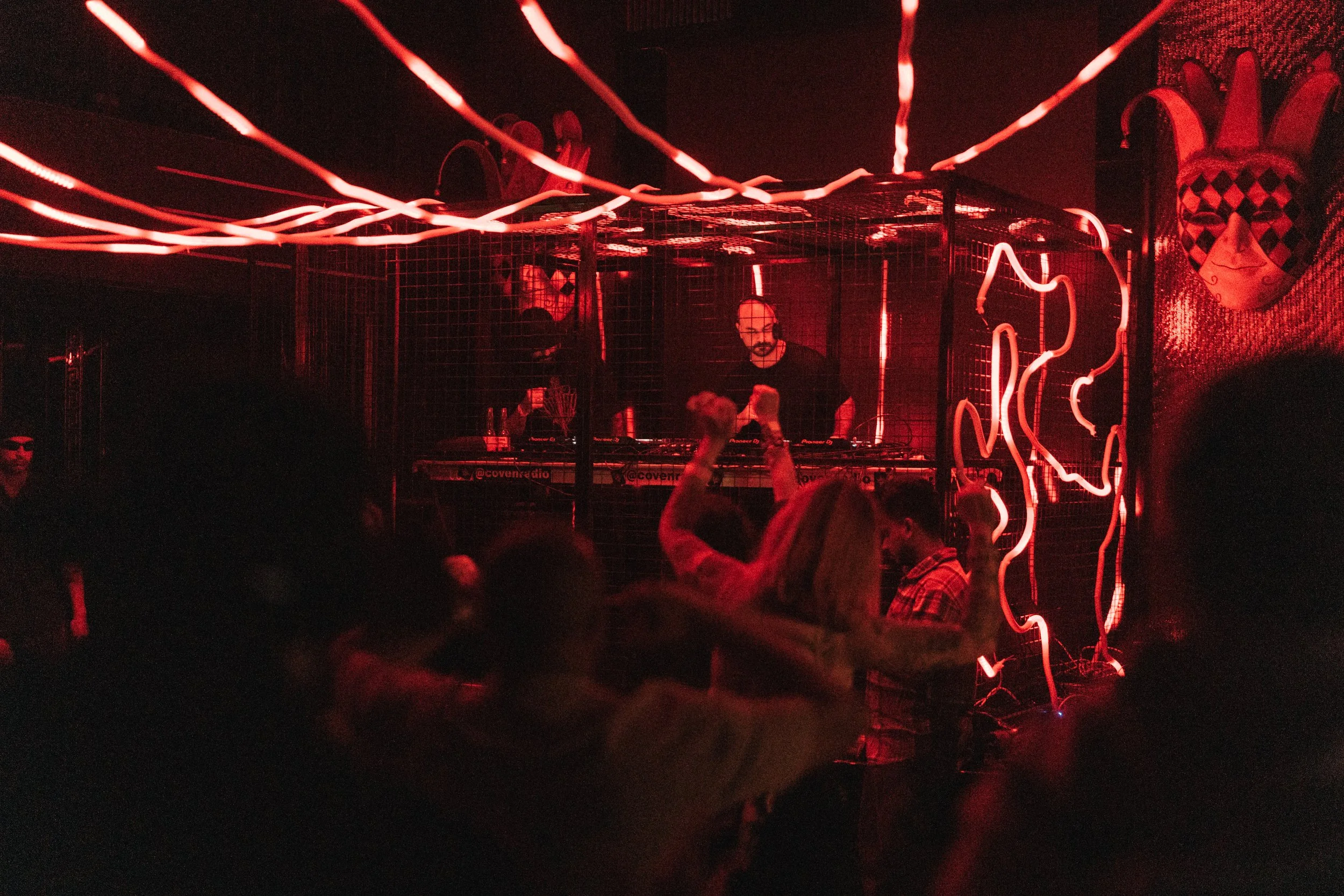 A nightclub scene with a DJ behind a booth in a cage, illuminated by red neon lights. People dance in front of the DJ, one with arms raised.