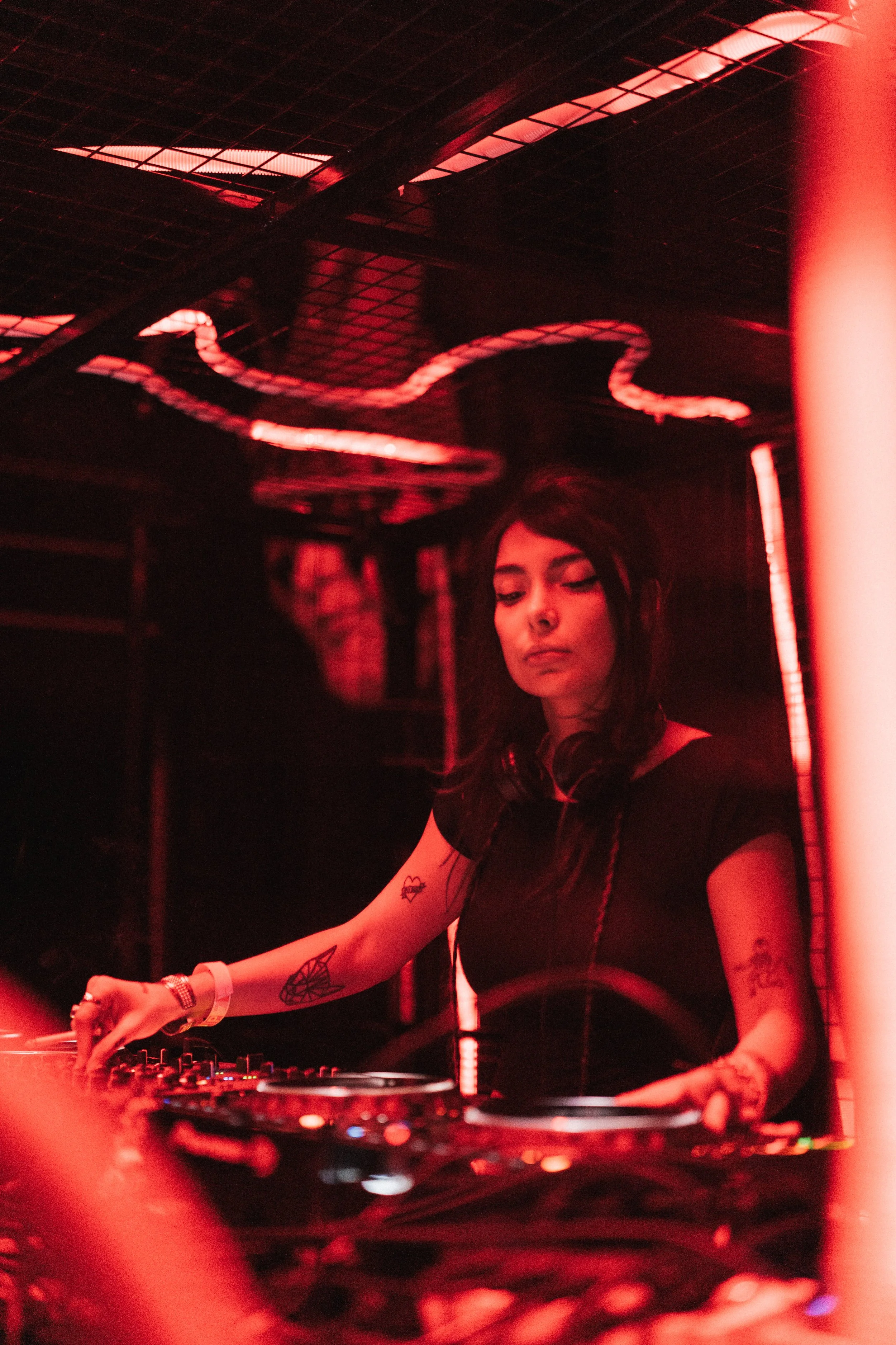A female DJ with tattoos on her arms, wearing headphones, mixing music at a club with red neon lighting.