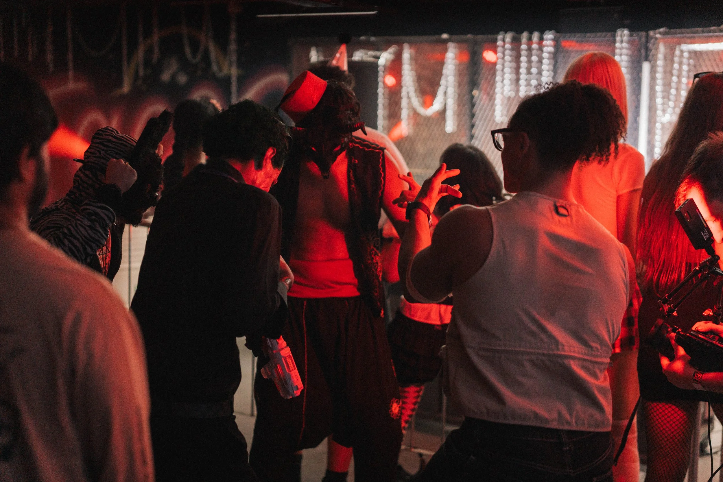 A group of people dancing and socializing at a party with red lighting, some wearing costumes and accessories, in an indoor venue with string lights and metal fencing in the background.