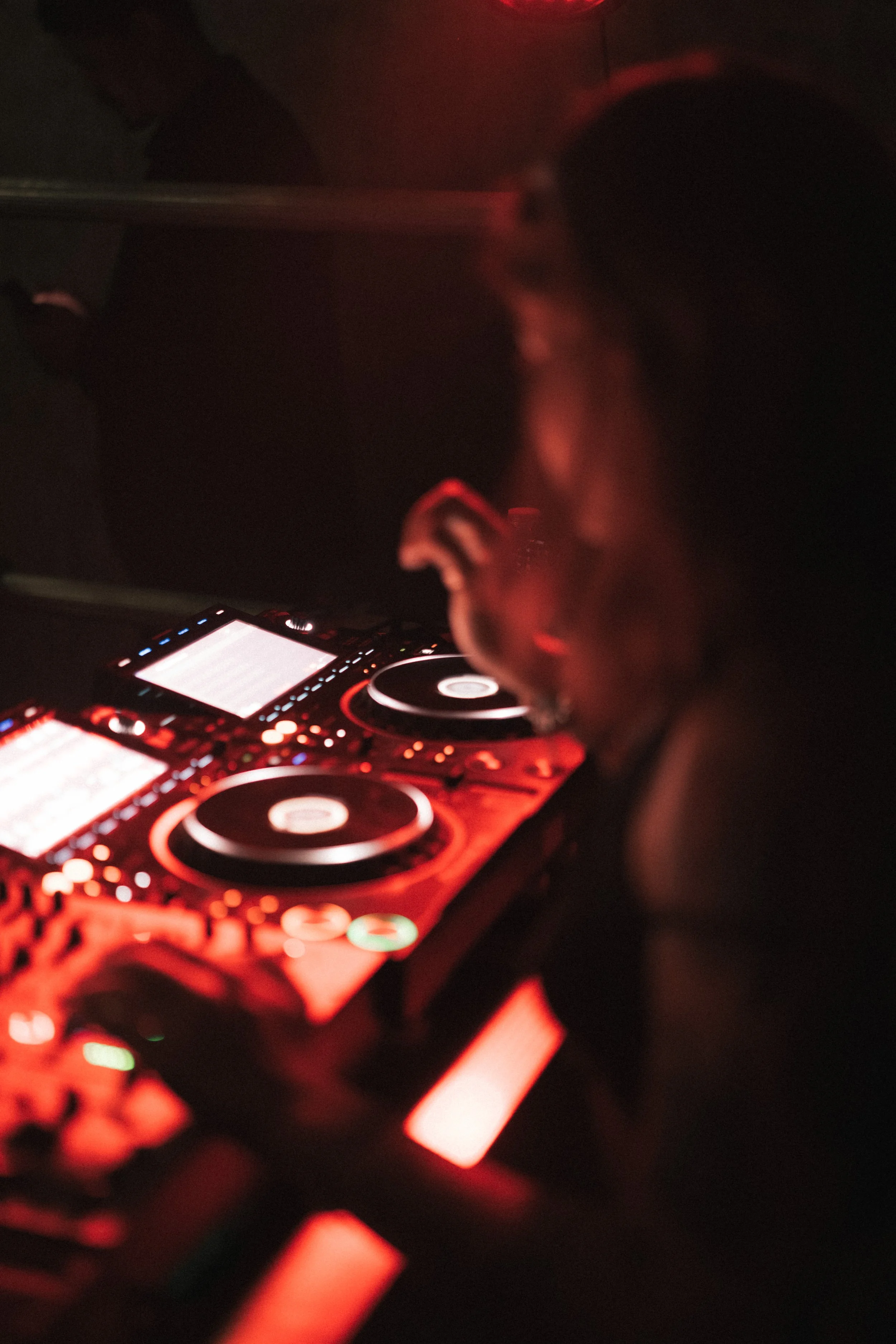 A DJ wearing headphones is mixing music on a DJ controller illuminated with red lights in a dark setting.