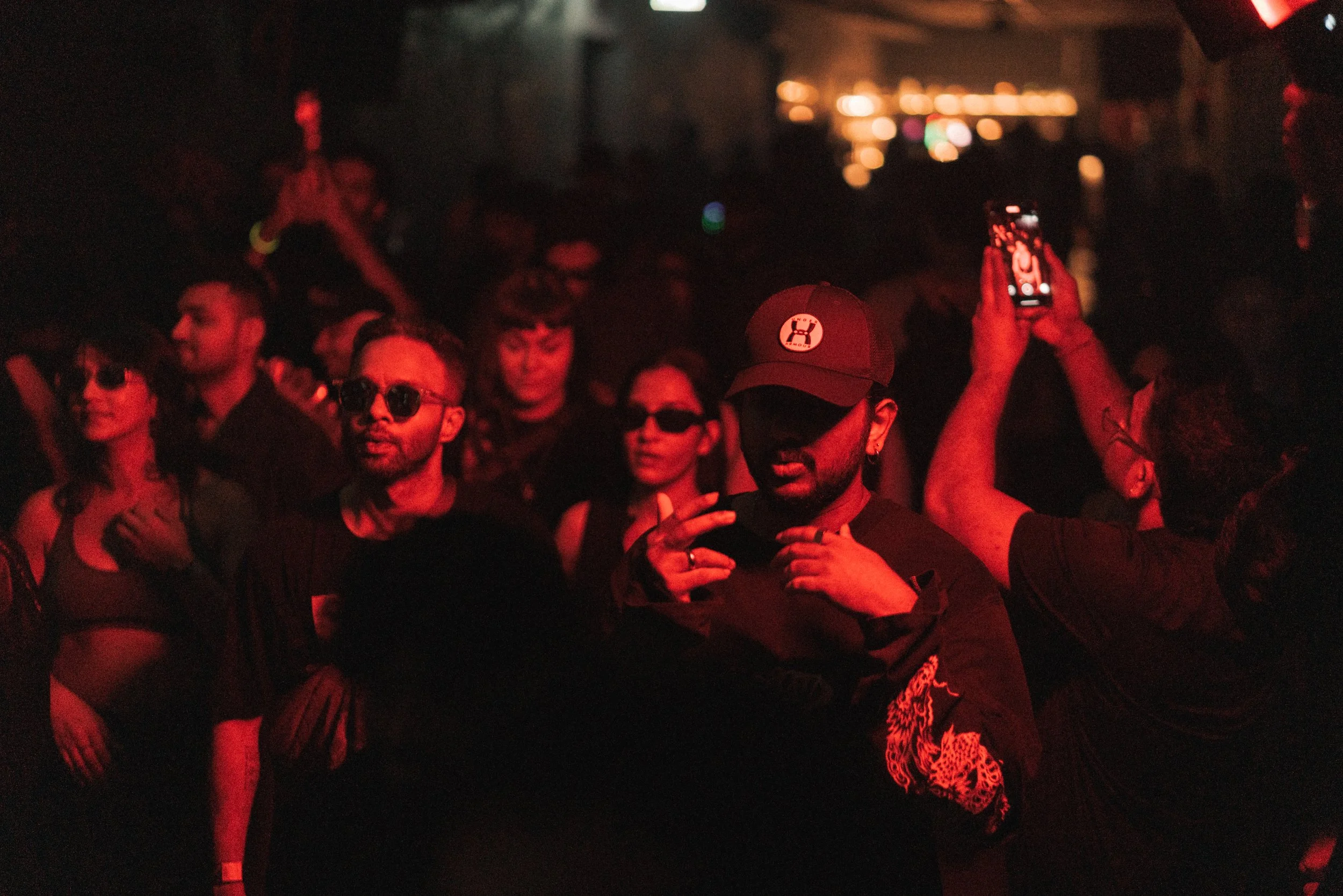 A group of people at a nighttime event, with red lighting and some wearing sunglasses; some are talking, and one person is taking a photo with a smartphone.